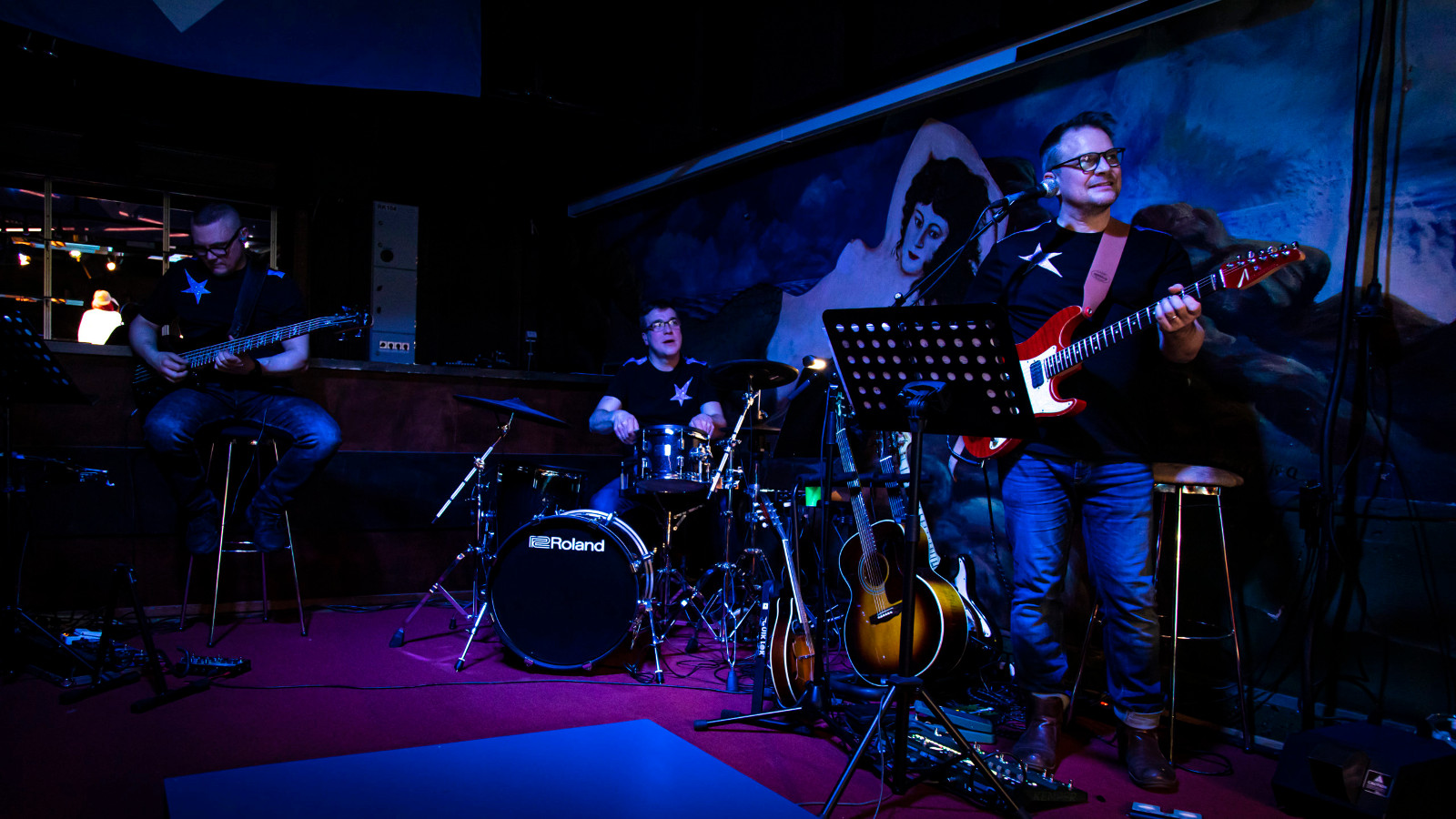 Kuvassa on bändi ja vasemmalla on kitaristi istumassa, vieressä rumpali ja oikealla basisti seisomassa. Kuvan sävy on tumma.