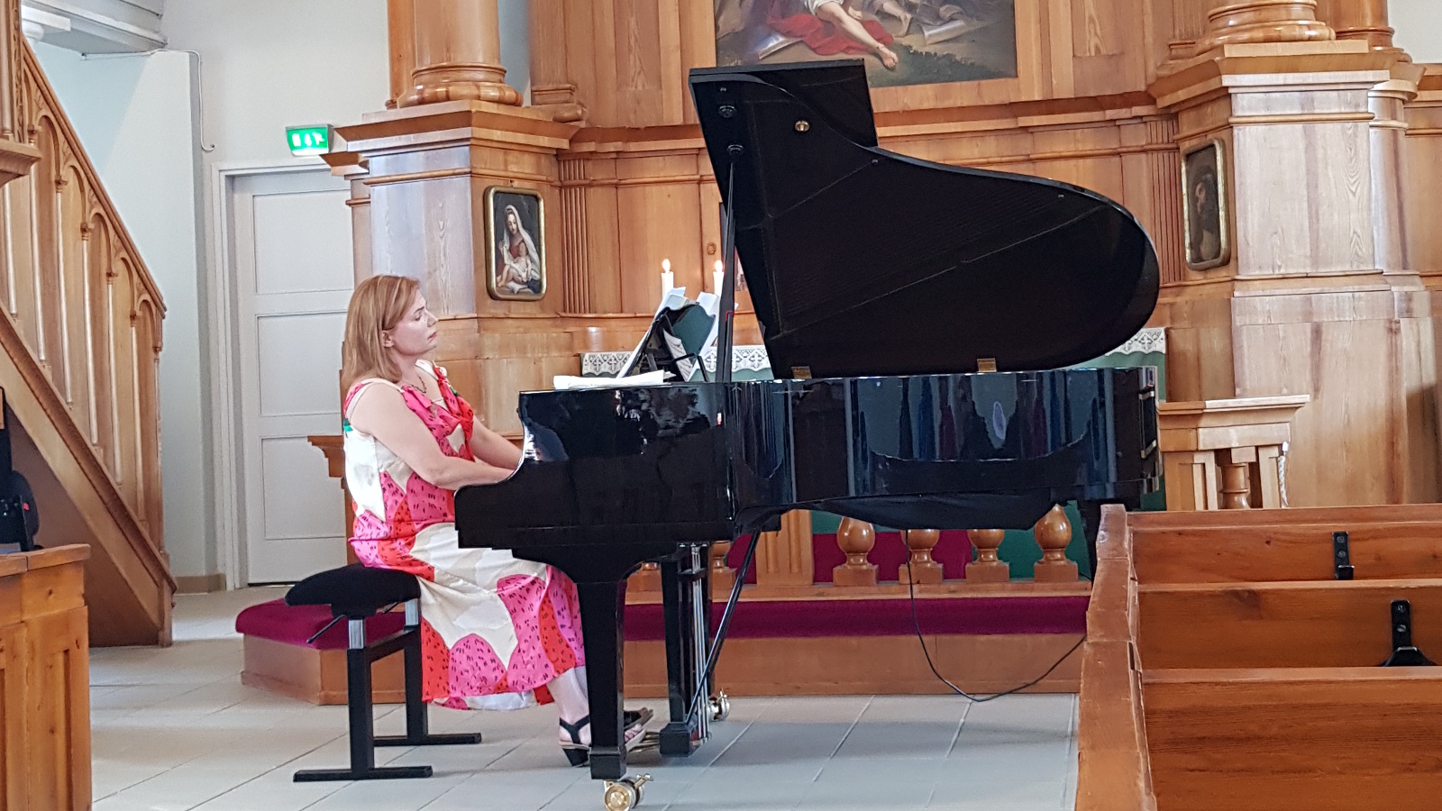 Kuvassa on Liisa Malmivaara soittamassa sivuttain Haminan kirkon kuorissa. Hänellä on vaalean ja punaisen kuviollinen mekko.