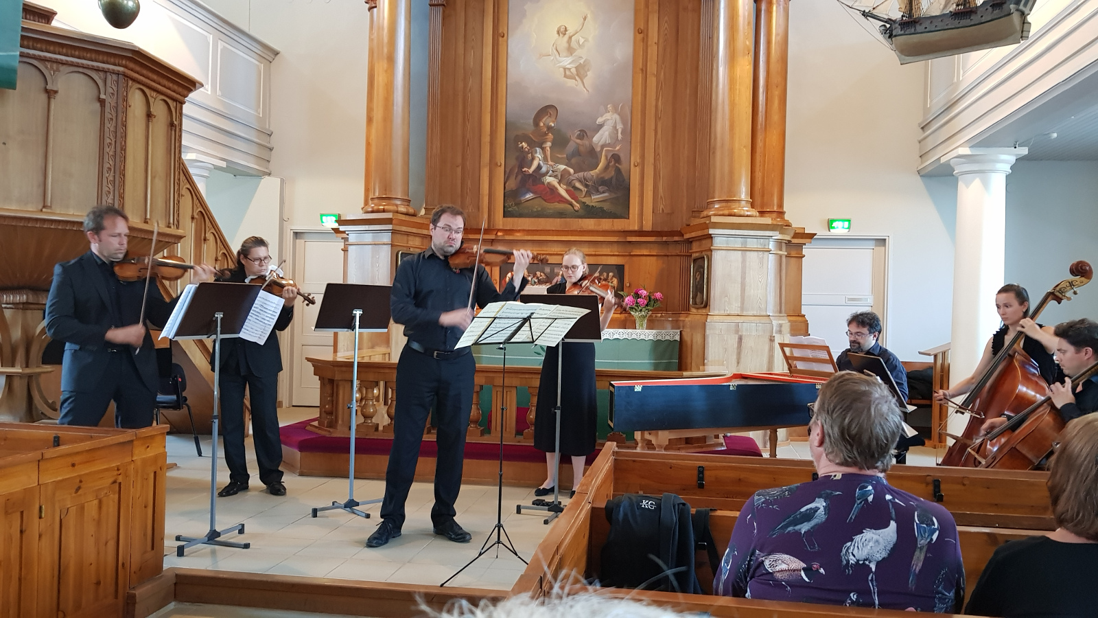 Kuvassa ovat Kaakon Kamarimusiikin ensemblen soittajat Haminan kirkon kuorissa koko kuvan leveydeltä. Taustalla näkyy alttaritaulua ja etualalla kirkon penkkejä sekä oikealla miehen selkämystä. 