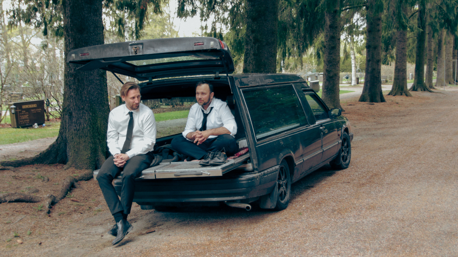 Kuvassa ovat Pekka Strang ja Jari Virman istumassa ruumisauton avonaisella takaluukulla. Auto on parkeerattu mäntykujalle hautausmaalla.