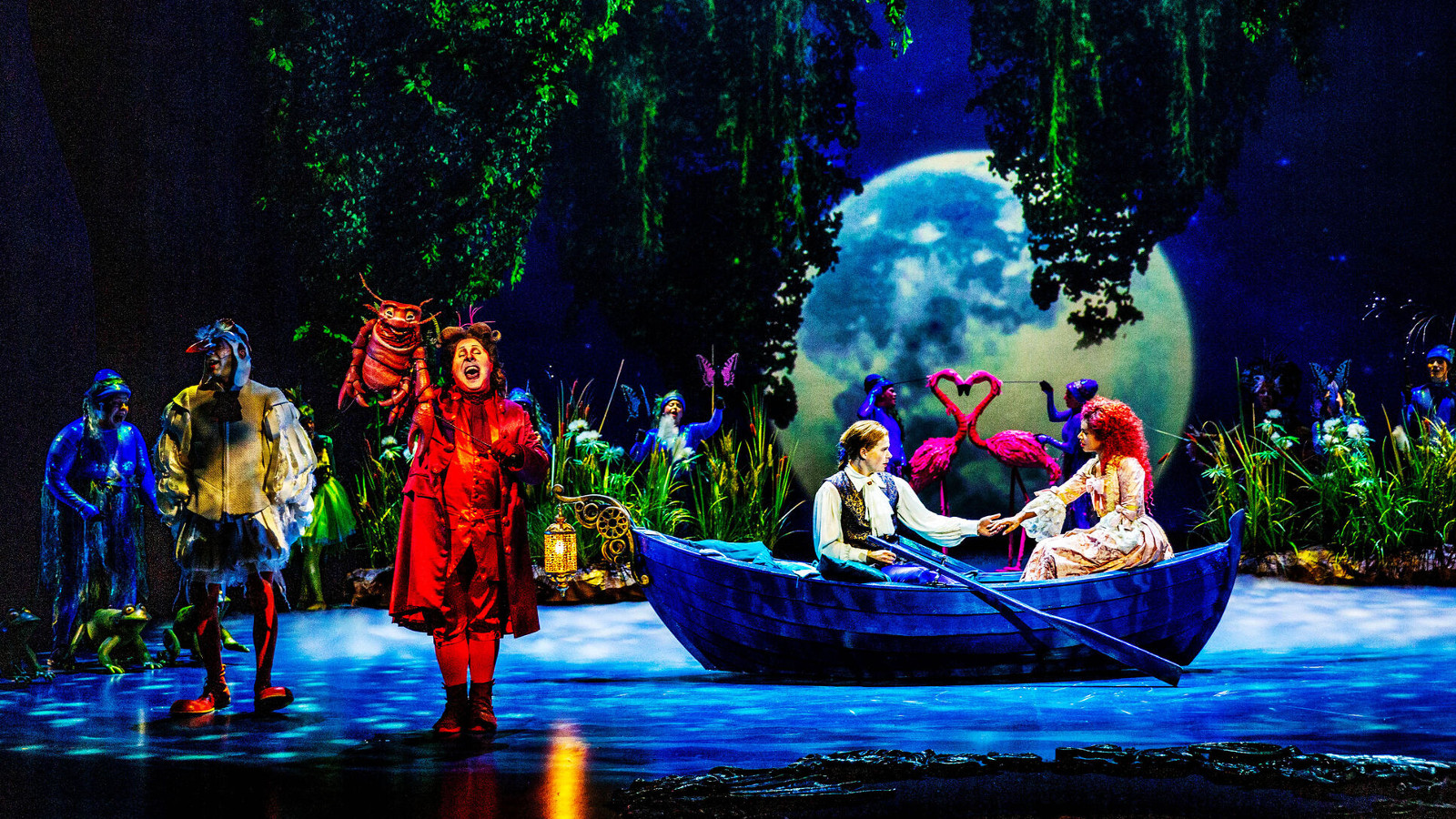 Kuvassa on lavalla on vasemmalla kolme hahmoa sinisissä ja punaisessa puvussa. Keskellä on sininen vene, jossa soutelevat tyttö ja poika. Taustalla on täysikuu ja ylhäältä roikkuu vihreitä oksia.