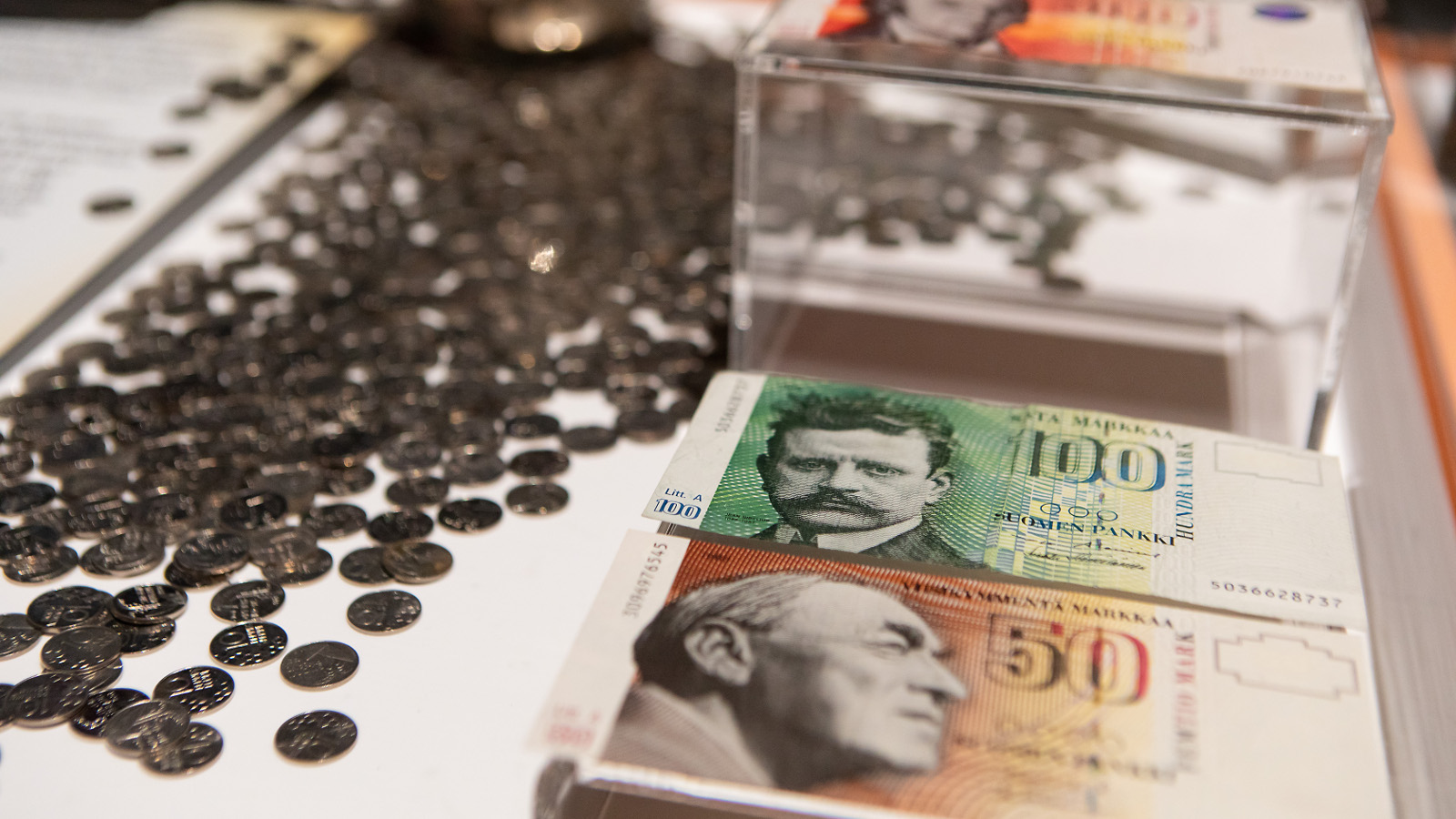 Kuvassa on vinosti aseteltuna oikealla ruskeita 50 markan, vihreitä 100 markan ja punaisia 500 markan seteleitä pinoissa. Vasemmalla on 10 pennin hopeisia kolikoita.