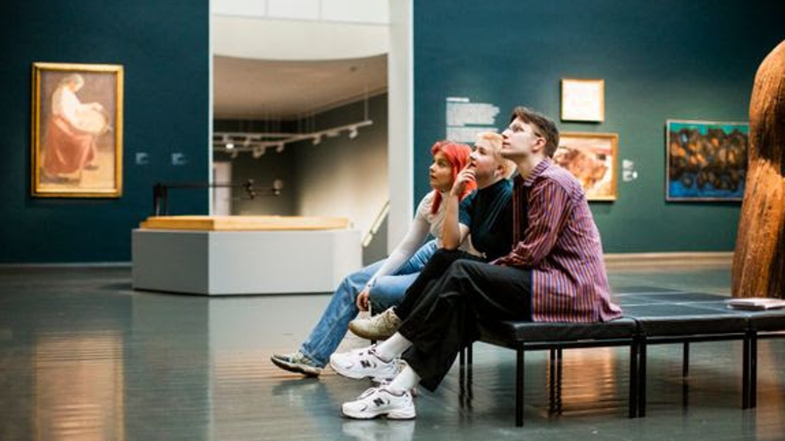 Kuvassa on kaksi nuorta naista ja yksi mies istumassa taidemuseon penkllä ja he ovat kuvassa sivuttain.  Vasemmalla taaempana on tumma seinä, jossa on naista maalaisvaatteessa oleva nainen.  Oviaukon toisella puolella on lisää tauluja.