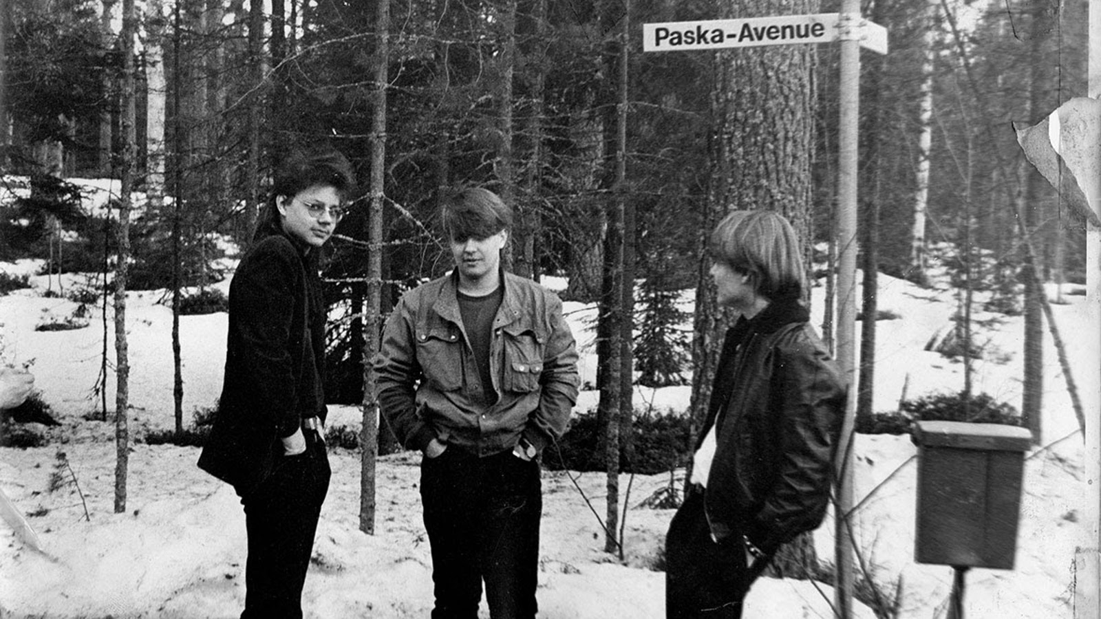Kuvassa on kolme poikaa, jotka seisovat metsikössä ja heidän luonaan on katukyltti, jossa lukee Paska-Avenue.  Heillä on pusakat ja mustat farkut päällään.