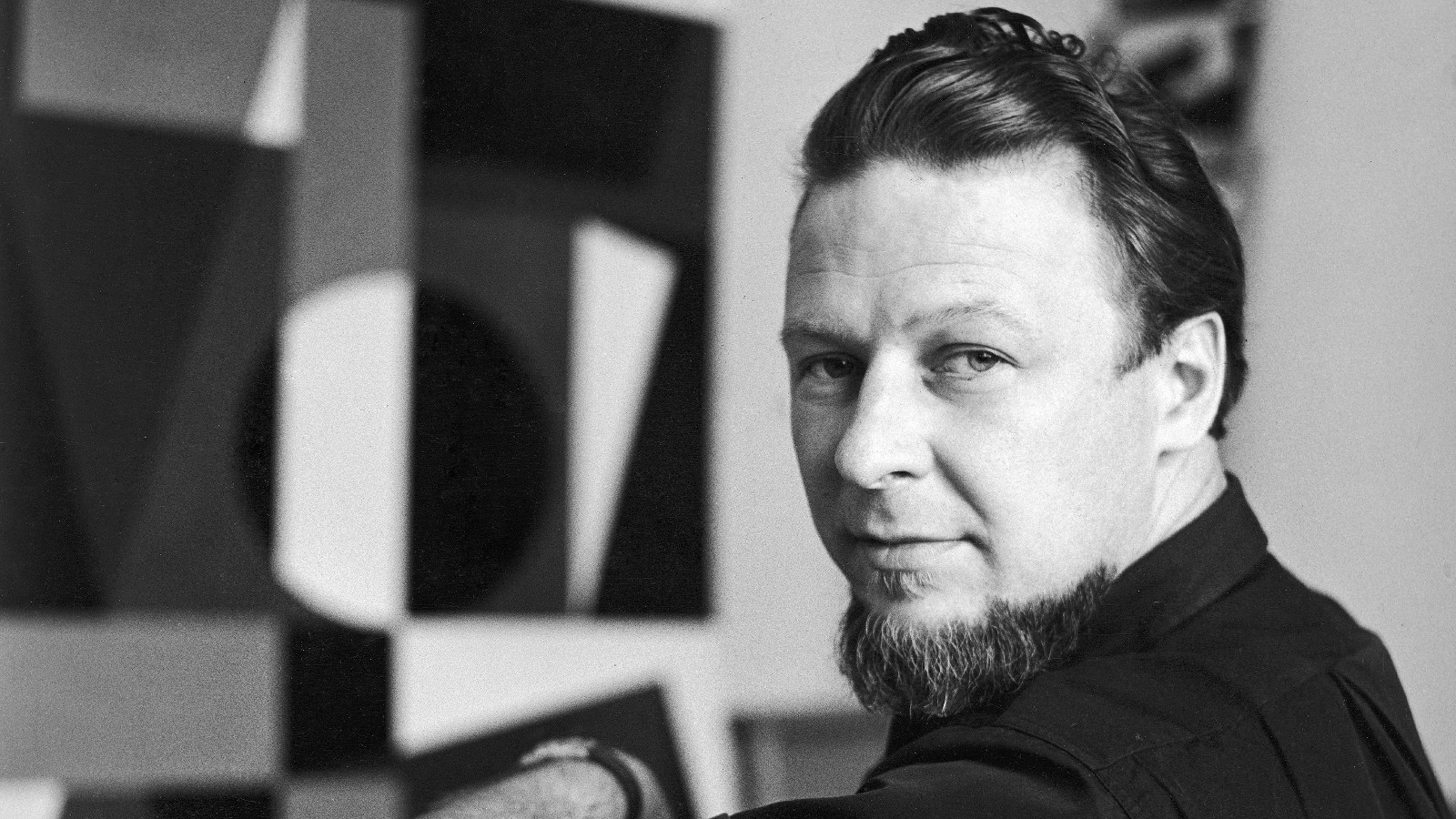 Kuvassa on Lars-Gunnar Nordström, joka istuu ja katsoo olkapäänsä yli. Taustalla näkyy taiteilijan teos epätarkasti. Kuva on musta-valkoinen.