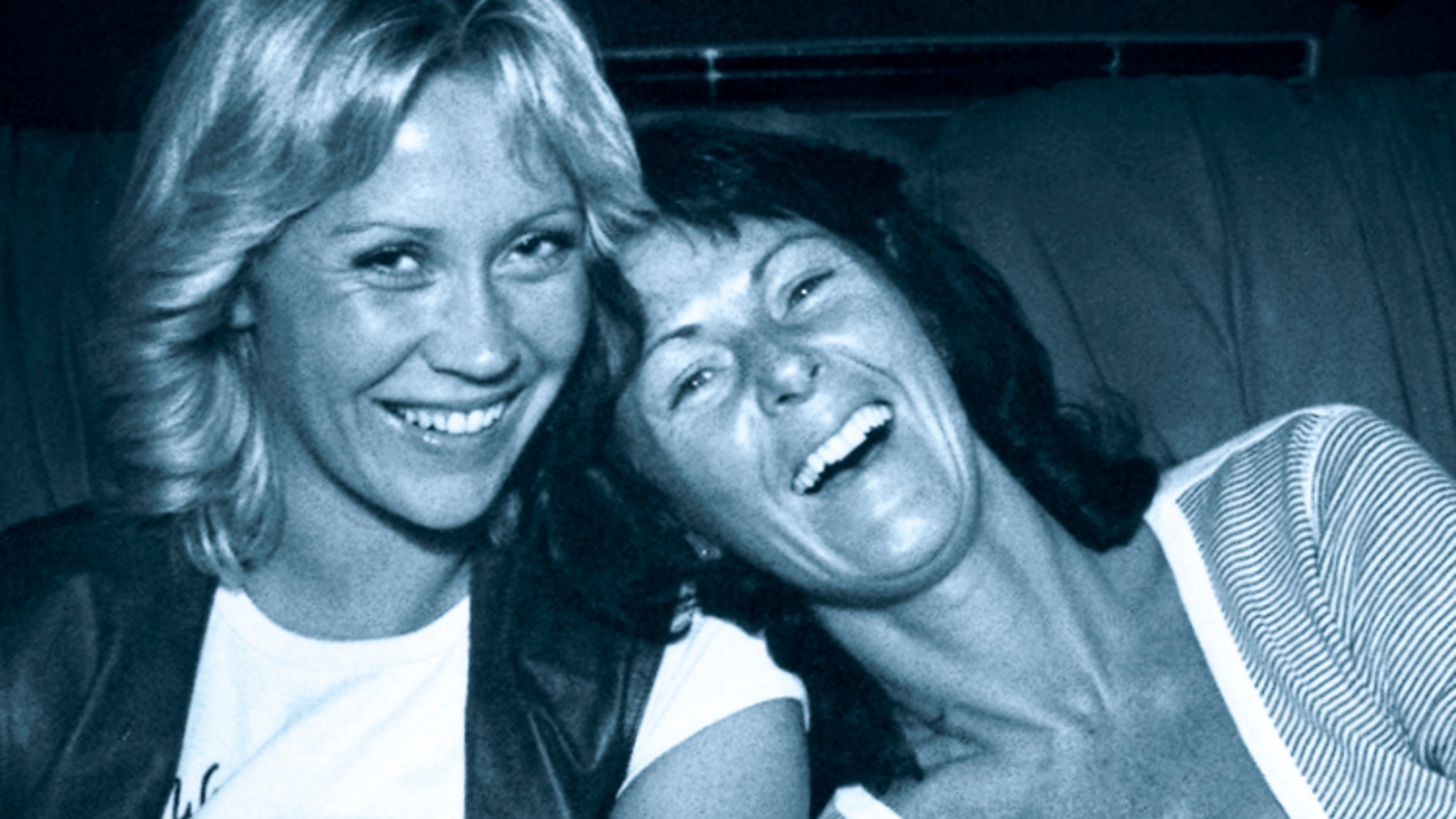 Kuvassa ovat Agneta Fältskog ja Ann-Frid Lyngstad kasvokuvassa.  Vasemmalla on vaaleatukkainen Fältskog ja häneen nojaa tummatukkainen Lyngstad. He molemmat nauravat.