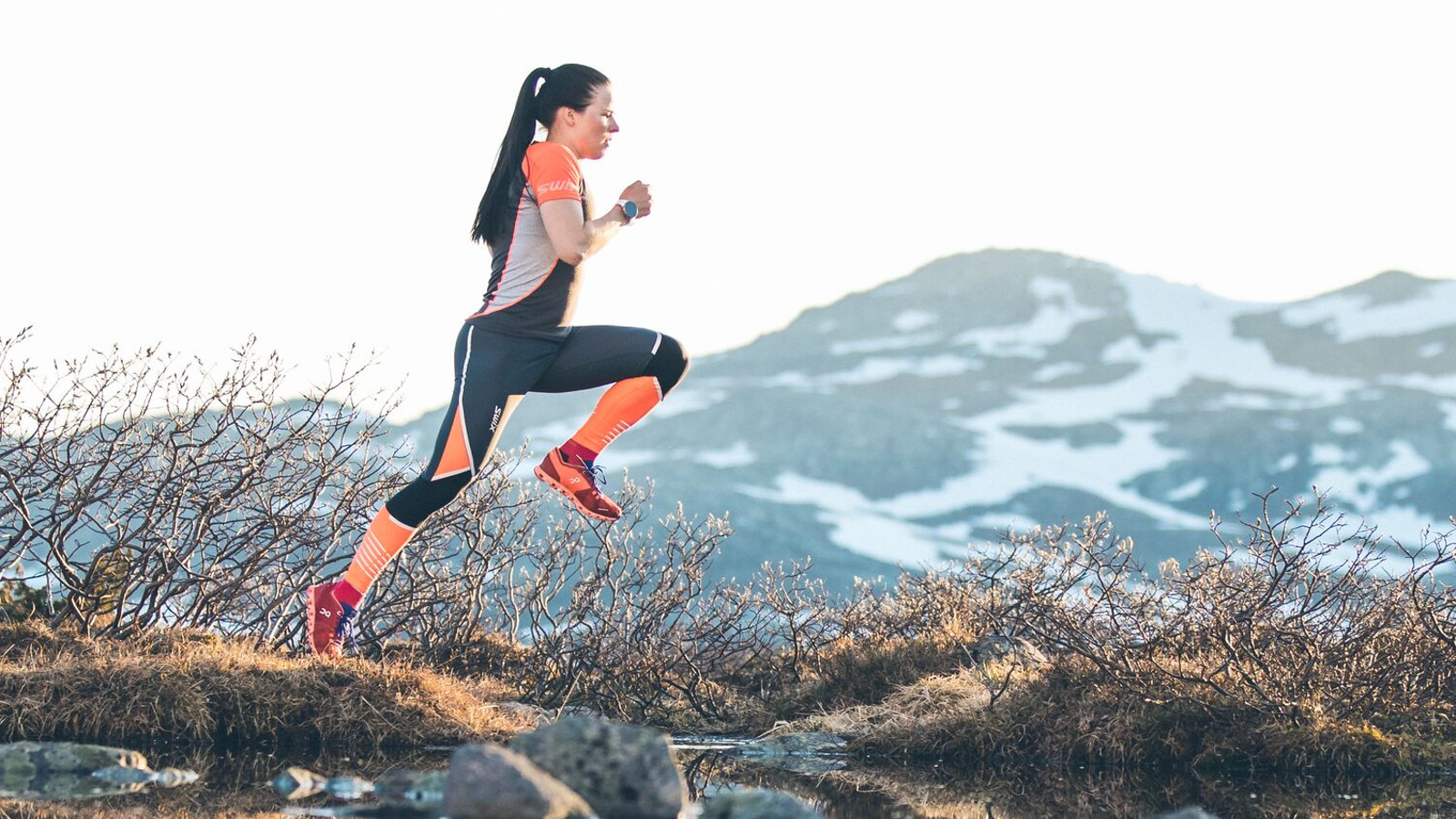 Kuvassa on Krista Pärmäkoski hyppäämässä vuoristossa hyppäämässä ojan yli. Takana on pensaita. Taustalla näkyy vuoristoa. Hänellä on musta-oranssi treeniasu.