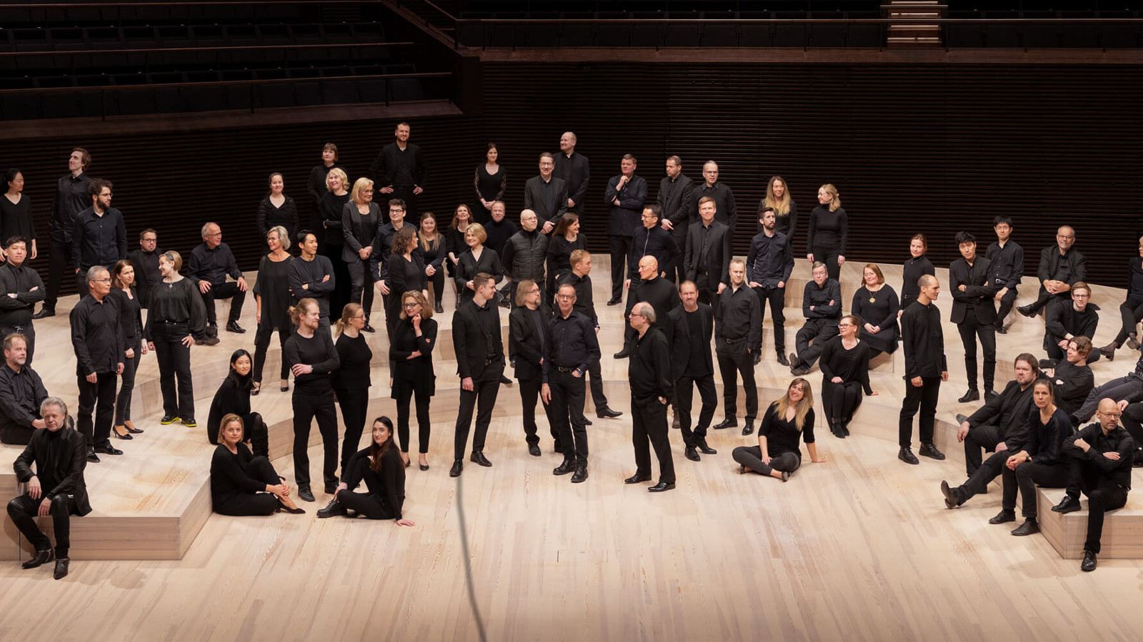 Kuvassa on RSO:n soittajia seisomassa mustissa vaatteissa sekalaisesti vaalean puisella esiintymiskorokkeella.