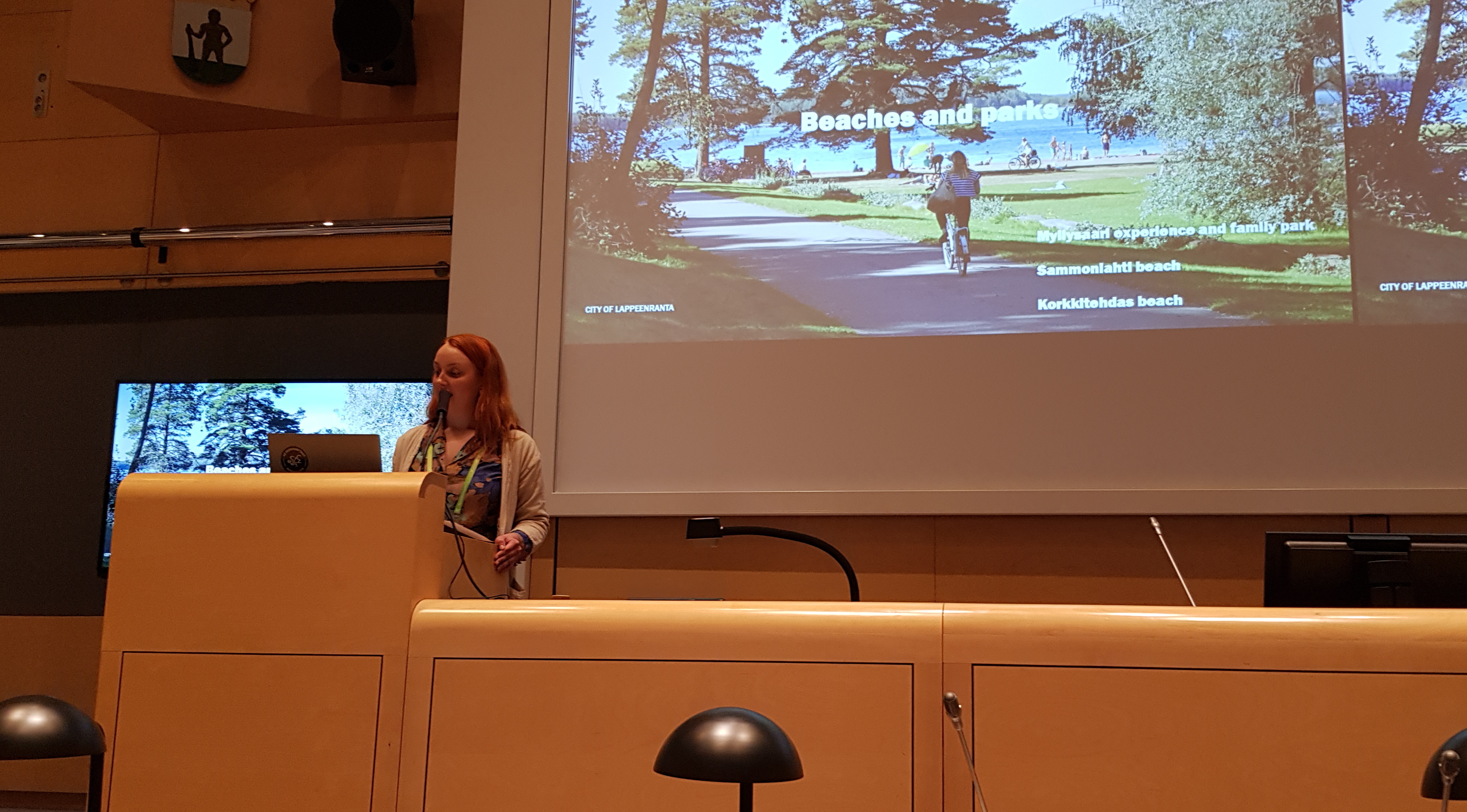 Kuvassa on Anita Pölönen puhumassa puhujapöntöllä ja vieressä näkyy puheenjohtajien pöytää.  Taustalla oikealla näkyy iso näyttö ja kesäistä puistomaisemaa, jossa kulkee kävelytie.  Taustalla puiden takaa näkyy sinistä järveä.  Vasemmalla näkyy osa pienestä näytöstä.