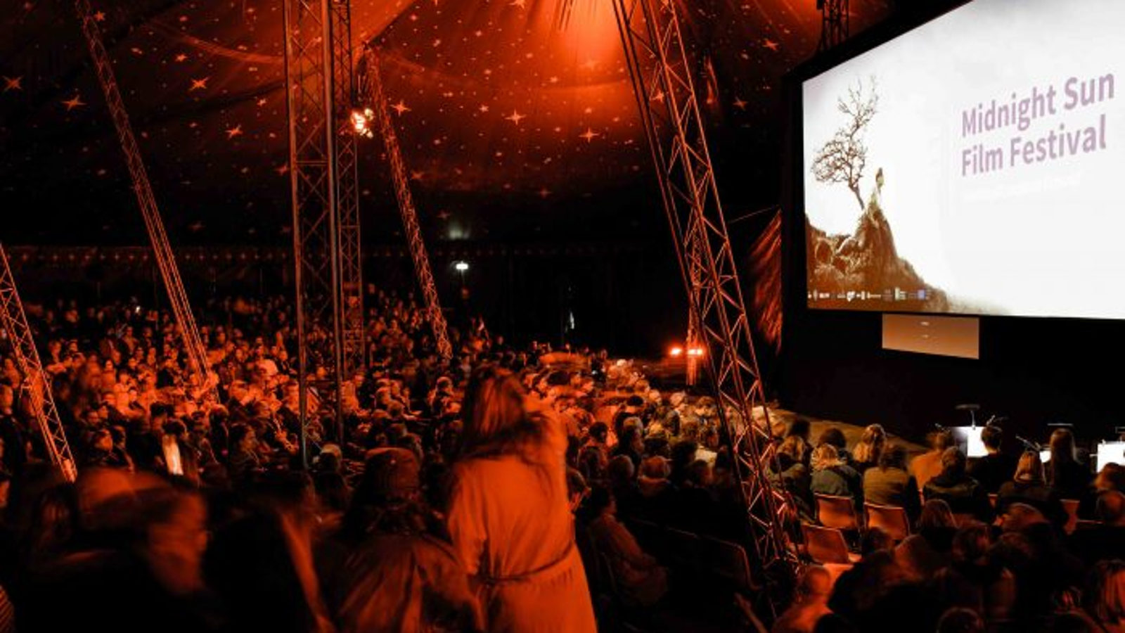 Kuva on Sodankylän elokuvajuhlilta ja pimennetyssä esitysteltassa on punaista valoa ja paljon yleisöä selin.  Meneillään on Moulin Rouge -karaoke.  Valkokankaalla lukee Midnight Sun Filmfestival.