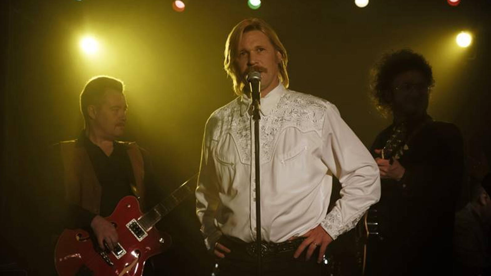 Kuvassa Kari Tapiota esittävä Matti Ristinen seisoo mikrofonin ääressä päällään valkoinen paita, jossa on yläosassa ja hihoissa kirjailua.  Vieressä on basisti soittamassa punaisella kitaralla ja taustalta näkyy keltavaloa tummalla taustalla sekä valkeita valoja ylä reunassa.