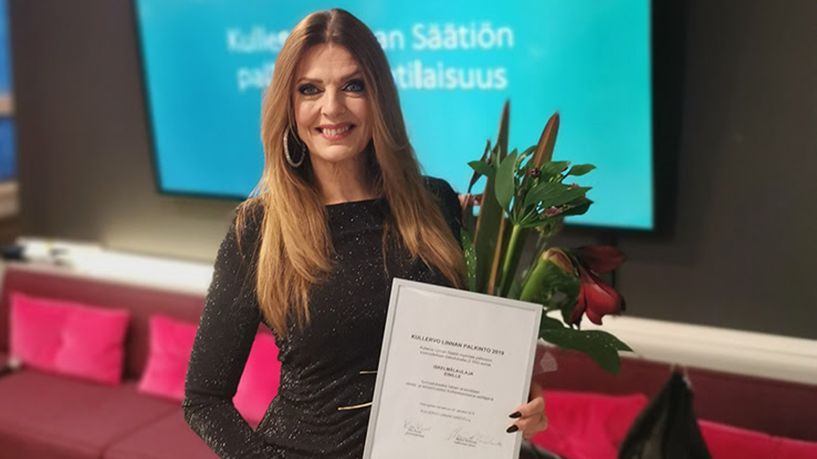 Kuvassa on Eini kädessään Kullero Linna -palkintotaulu ja onnittelukukat. 