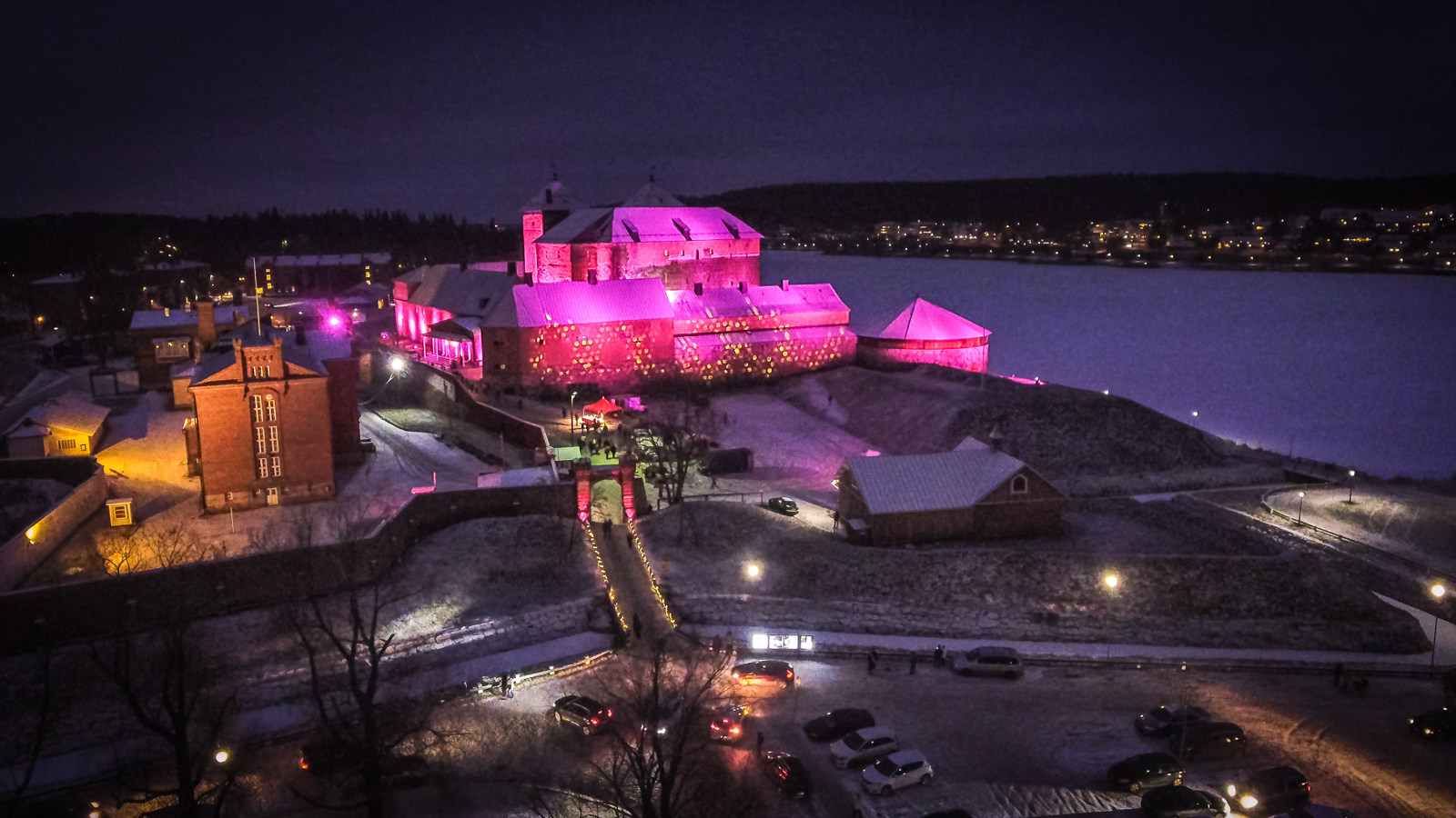 Kuvassa on Hämeen linna valaistuna punertavaksi ympäristössään.  Kuva on otettu illalla ja pimeällä.
