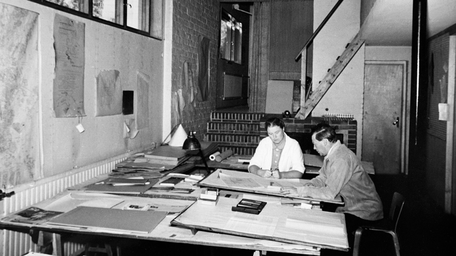 Kuvassa ovat Aino ja Alvar Aalto työskentelemässä arkkitehtitoimistossaan.  Huone on tilava ja työskentelytila oli alhaalla, jossa pariskunta työskenteli iso pöydän ääressä. Kuva o musta-valkoinen.