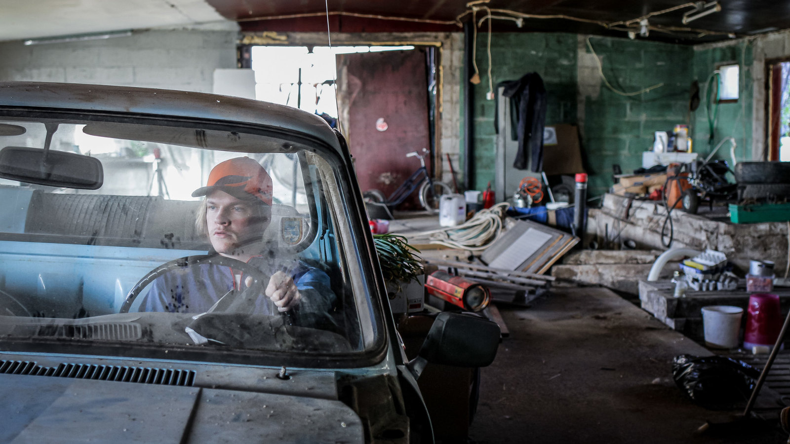 Kuvassa on Juho Uusitalo auton etuistuimella ja auto on autokorjaamolla.  Taustalla näkyy erilaista autokorjaamotavaraa.