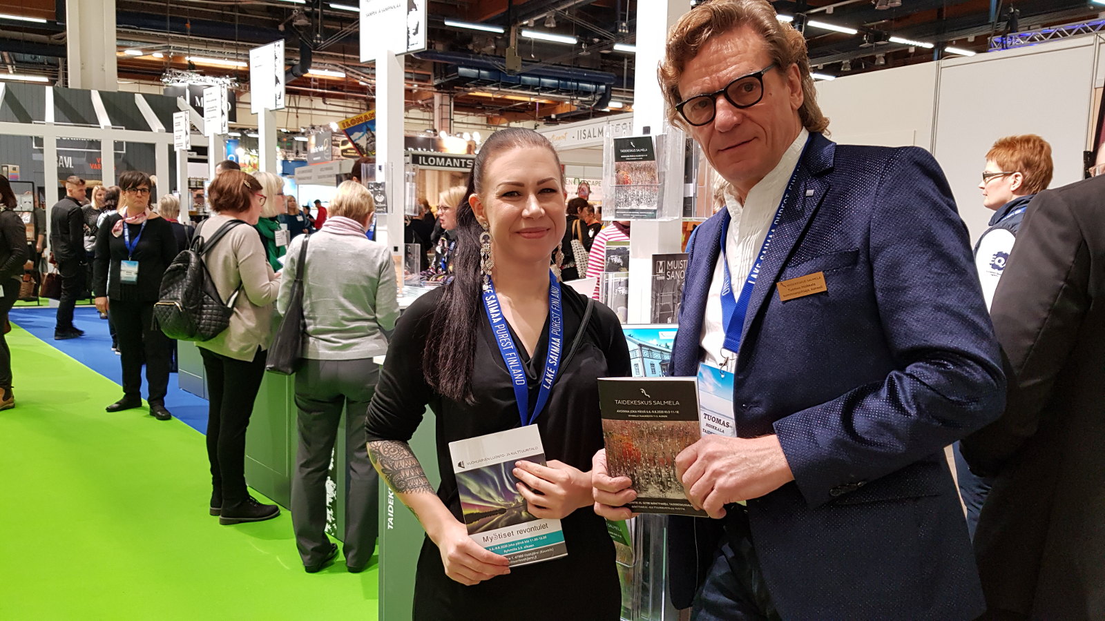 Kuvassa ovat Heidi Tuovinen ja Tuomas Hoikkala edustamassa Taidekeskus Salmelaa  Helsingin matkamessuilla.  Heillä on kädessää esitteet ja taustalla näkyy vihreää mattoa ja muiden osallistujien esittelijöitä.