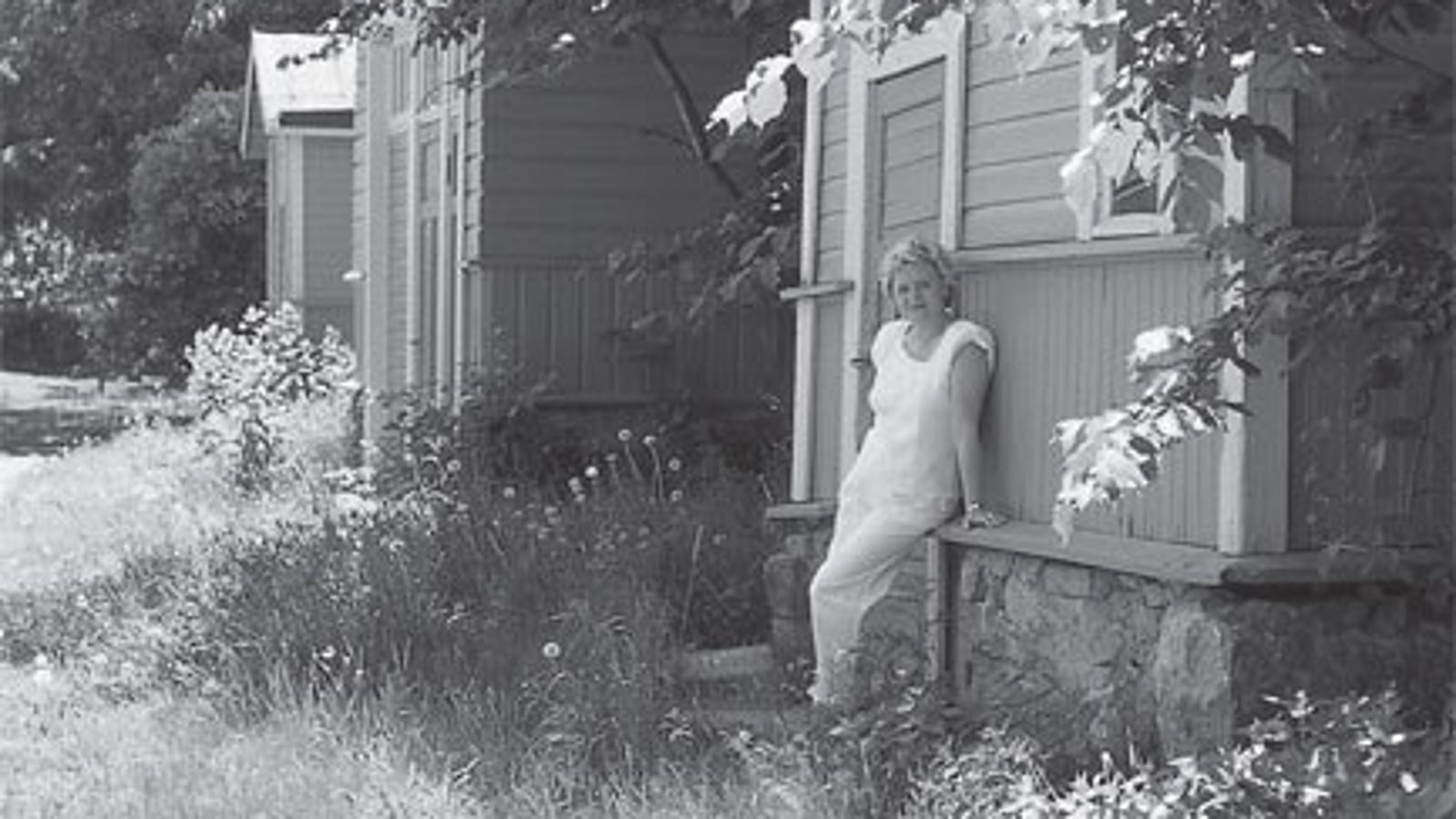 Kuvassa on Laila Hirvisaari Lappeenrannan linnoituksen puutalon pihalla.  Kuva on musta-valkoinen.