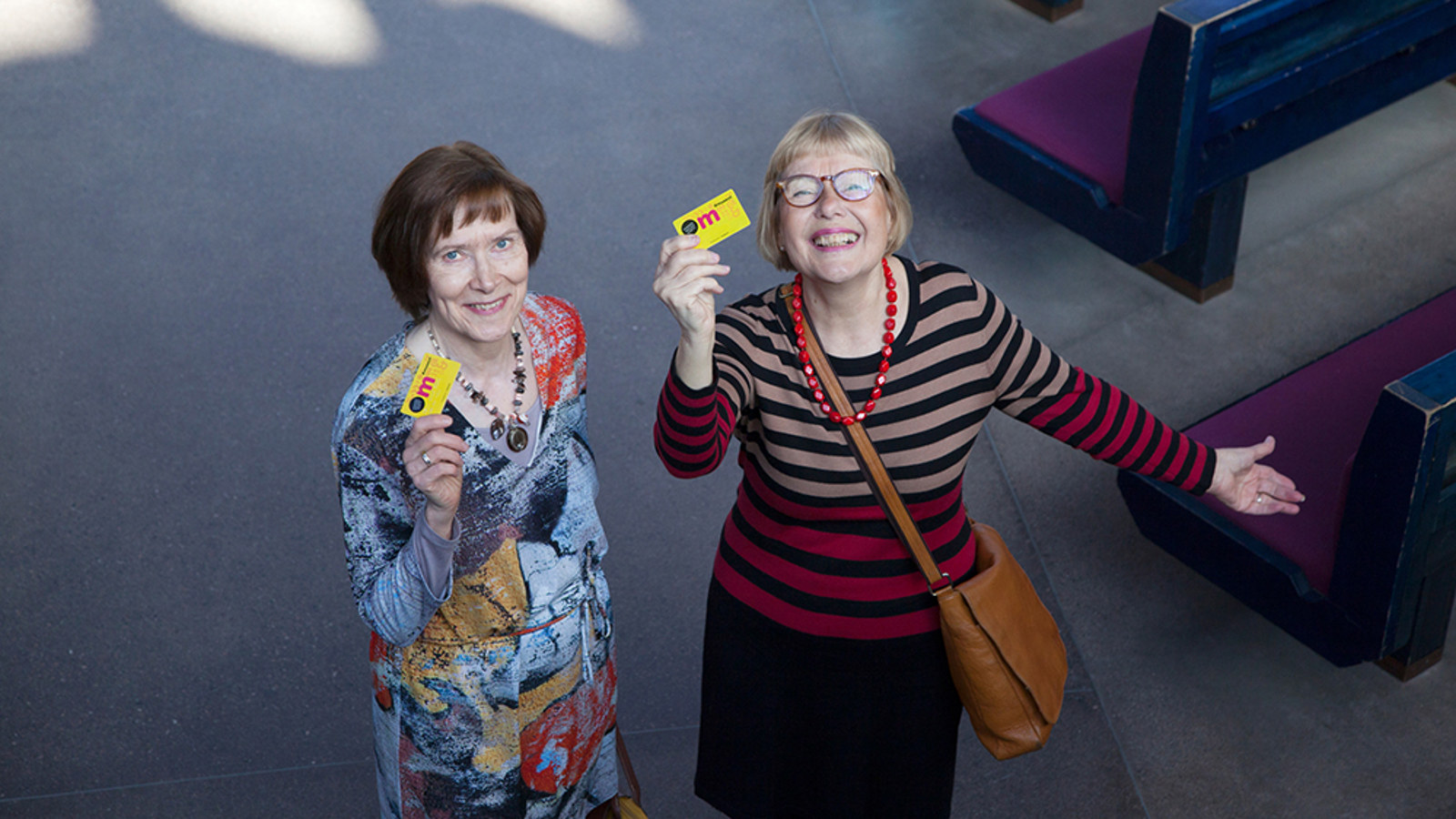 Kuvassa on kaksi keski-ikäistä naista kuvattuna ylhäältä päin.  Oikealla puolella olevalla on oikeassa kädessään museokortti, jota hän iloisesti näyttää. 