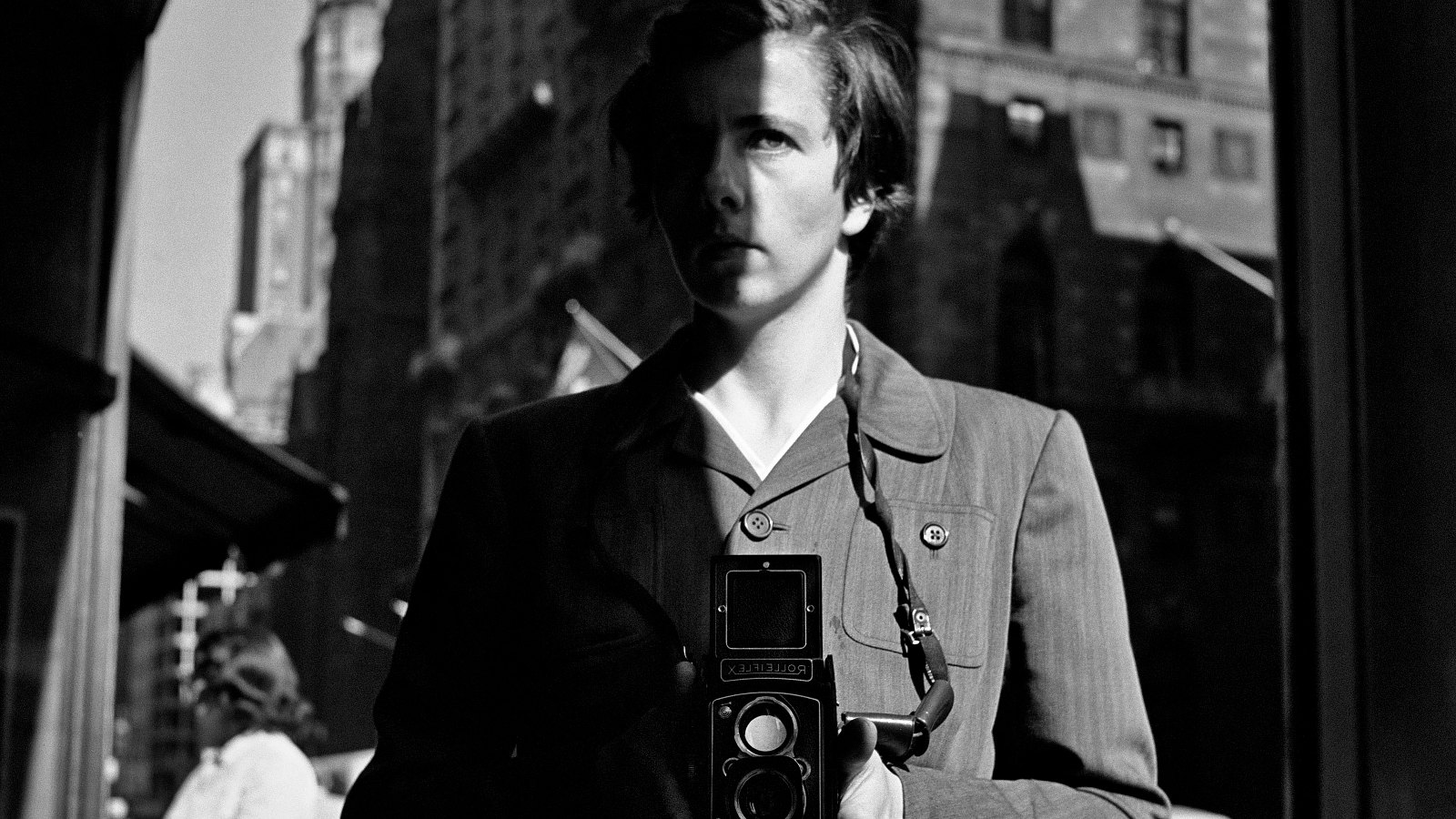 Kuvassa on Vivian Maier kamera kädessään vyötärön alapuolella.  Kuva on otettu New Yorkissa kadulla ja se on msuta-valkoinen.