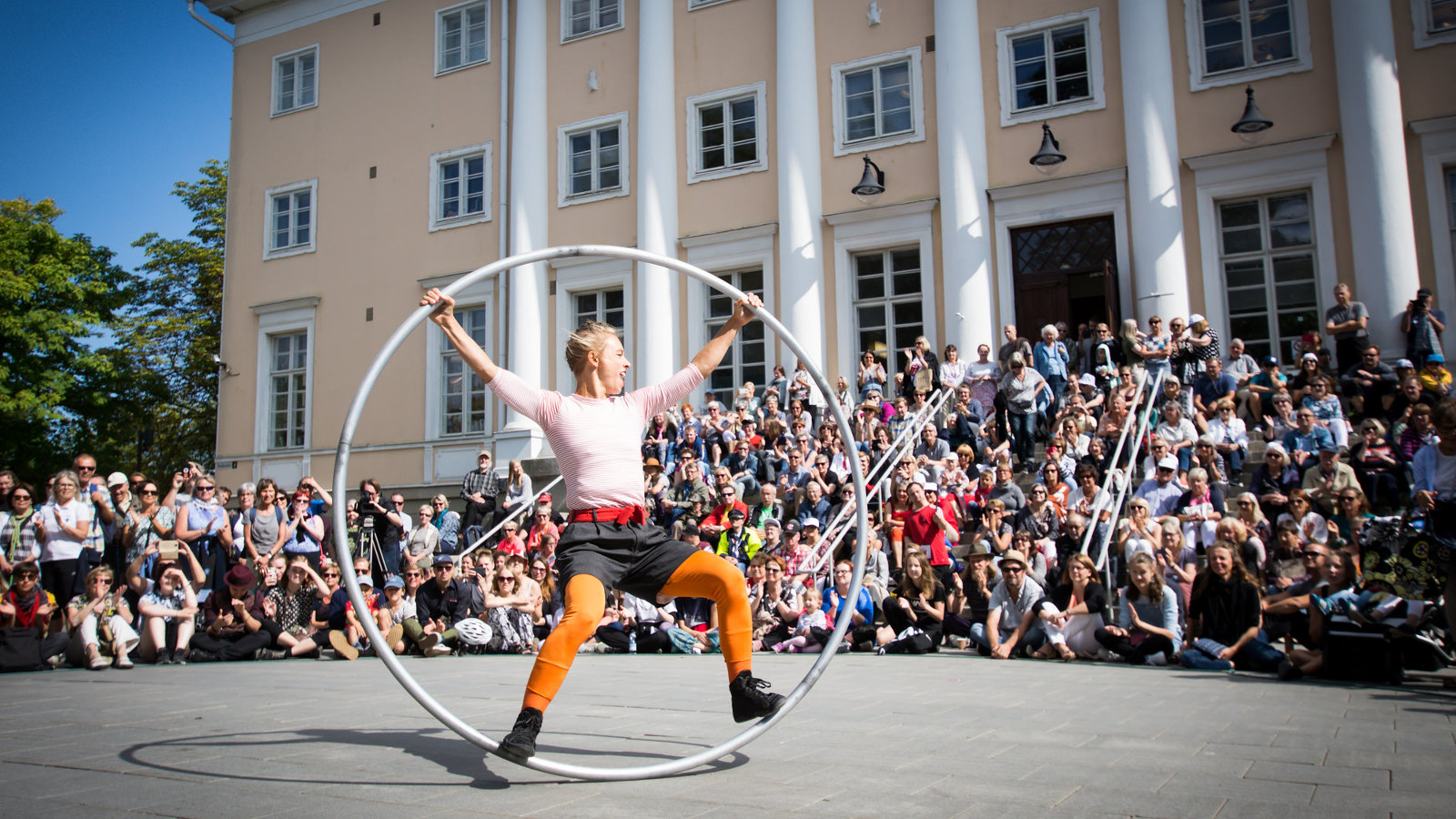 Kuvassa on Tampereen Teatterikesän esitys kadulla, jossa yleisöä keltaisen rakennuksen edessä.  Ilma on aurinkoisen kesäinen.