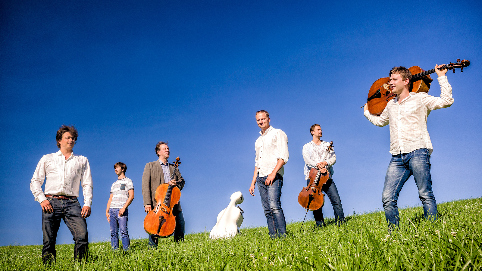 Kuvassa on Total Cello Ensemble, jonka kuusi jäsentä seisovat nurmikolla rinteen päällä.  Takana on kirkkaan sininen taivas.  Jäsenillä on valkoiset paidat ja farkut sekä soittimet mukanaan.