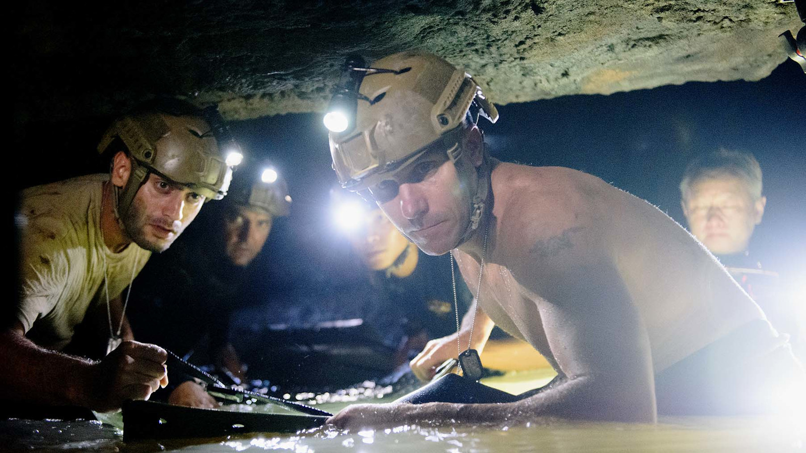 Kuvassa on kaksi pelastajaa luolassa keltaisissa kypärissä.  Vesi on lähes vyötäröä myöten ja luola on matala.  Miehet eivät voi seisoa suorassa.
