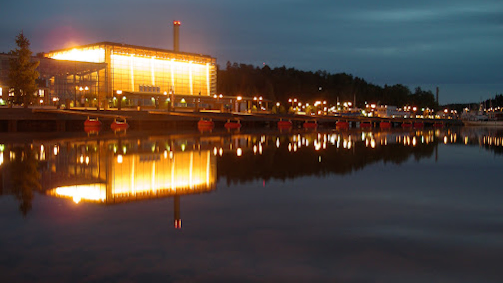 Kuvassa on Lahden Sibeliustalo iltavalaistuksessa ja kuva on otettu Vesijärven suunnasta.  Valot heijastuvat tummaan veteen.