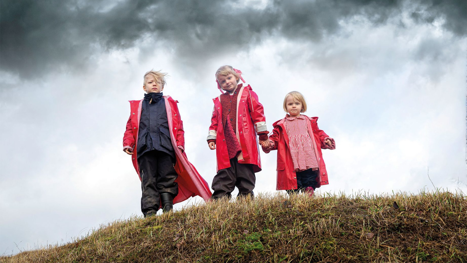 Kuvassa on kolme lasta kumpareen päällä ja heillä on päällään punaiset takit.