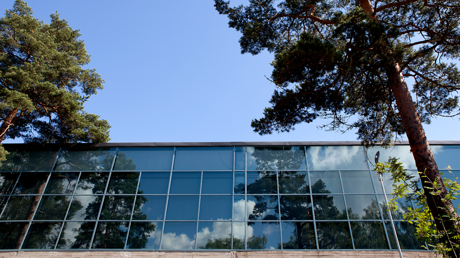 Kuvassa on EMMA taidemuseon lasinen julkisivu yläviistosti.  Taustalla on sininen taivas ja mäntyjä.