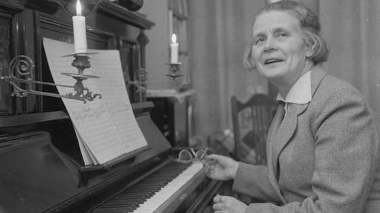 Kuvassa on Helvi Leiviskä vanhanaikaisen pianon ääressä pukeutuneena jakkuun.  Kuva on musta-valkoinen.