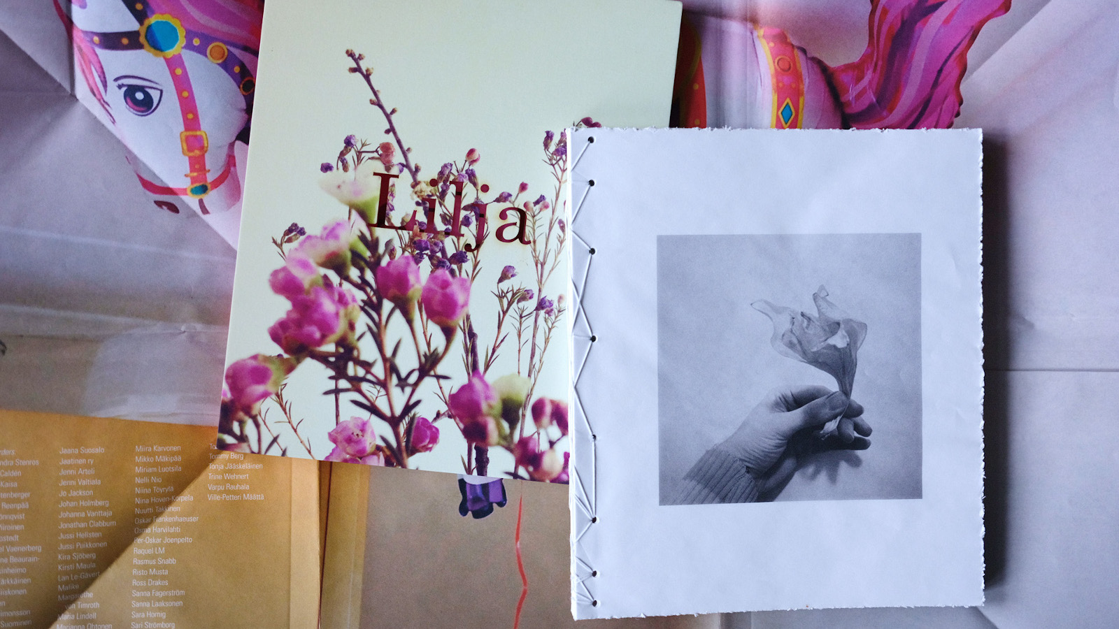 Kuvassa on kirjan Lilja kansi ja toisena on sisäsivu, jossa on on kuva kukasta. Taustalla on paperitaitoksia ja kuva pinkin punaisesta leikkihevosesta.