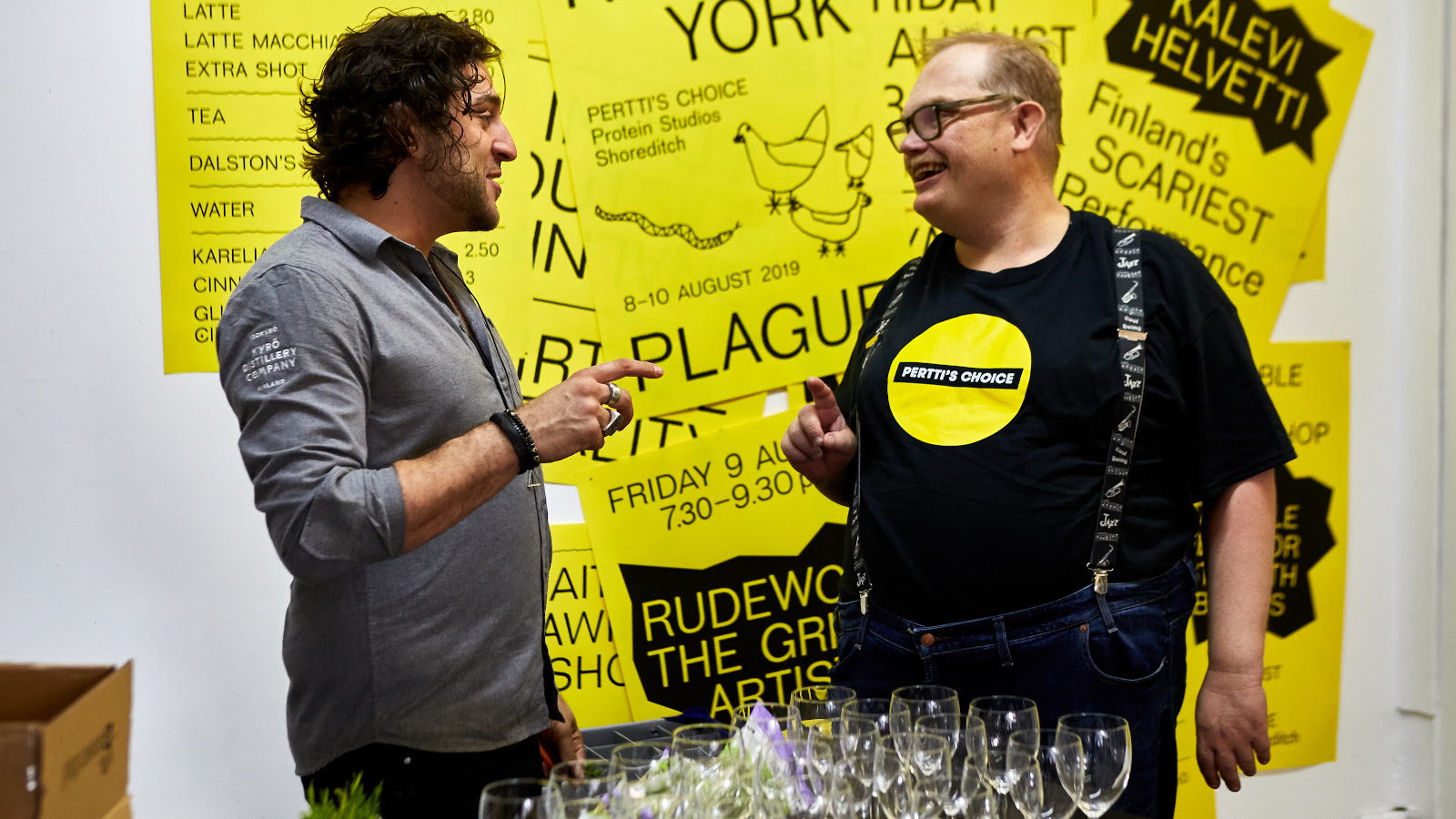 Kuvassa on Pertin Valinnan Sami Helle oikealla ja Kyrö Distilleryn edustaja kesksutelemassa edessään pöytä, jossa on laseja. Takana on keltaisella pohjalla julisteita mustalla tekstillä.