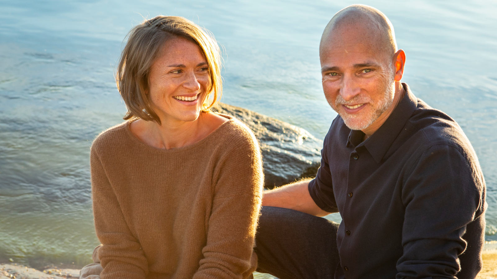 Kuvassa ovat Inka Kallen ja Aki Louhimies istumassa auringoisella ilmalla rantakalliolla meren rannalla.