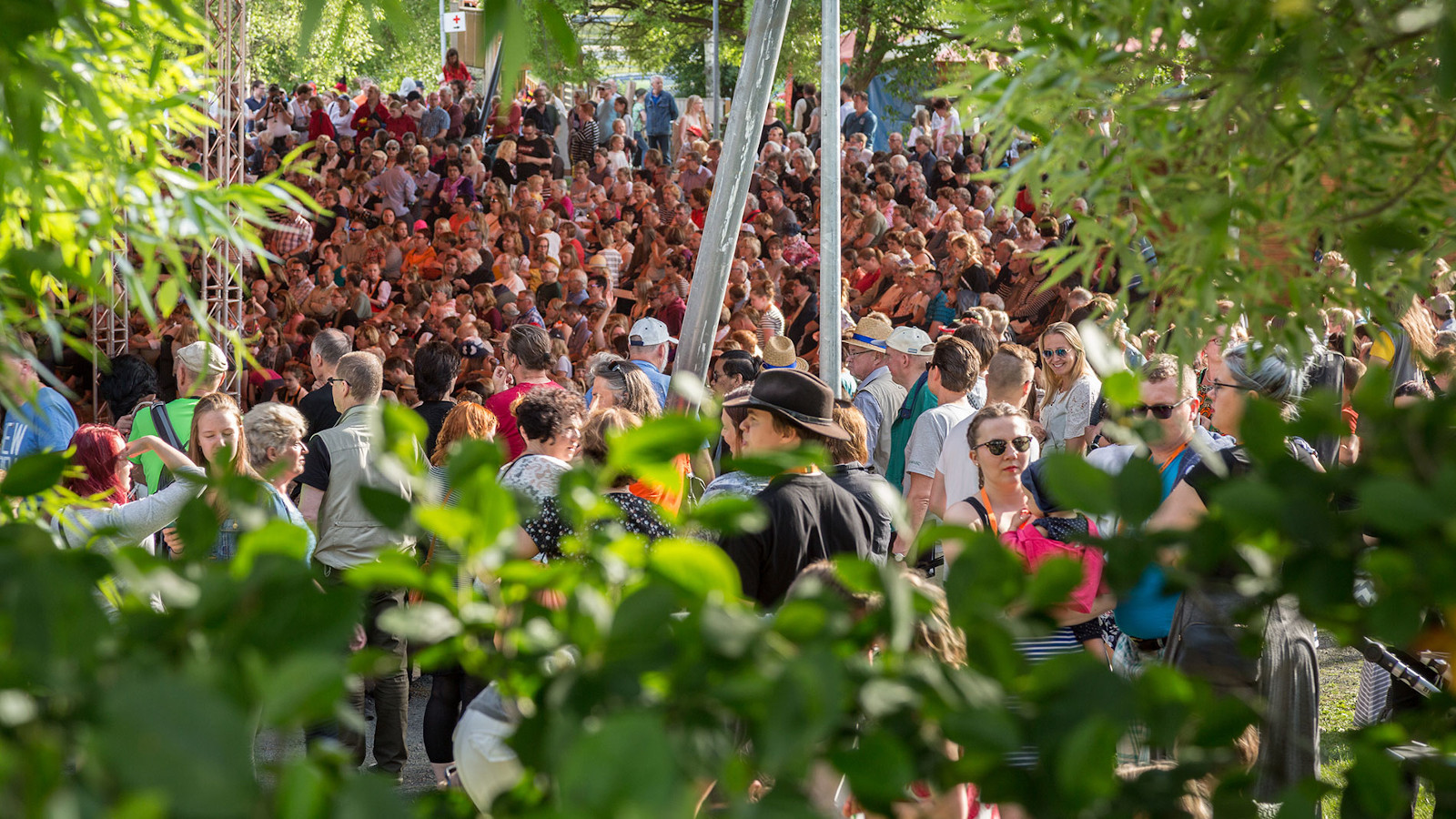 Kuvassa on Kaustisen kansanmusiikkijuhlien yleisöä, jotka ovat tiiviinä ryhmänä vihreiden puiden ympäröimänä.