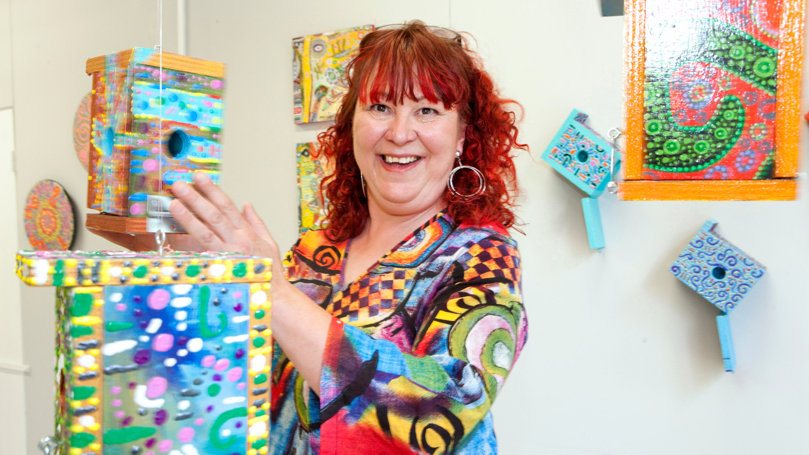 Kuvassa on Paula Huhtanen värikkäissä vaatteissa ja värikkäiden taideteostensa keskellä.