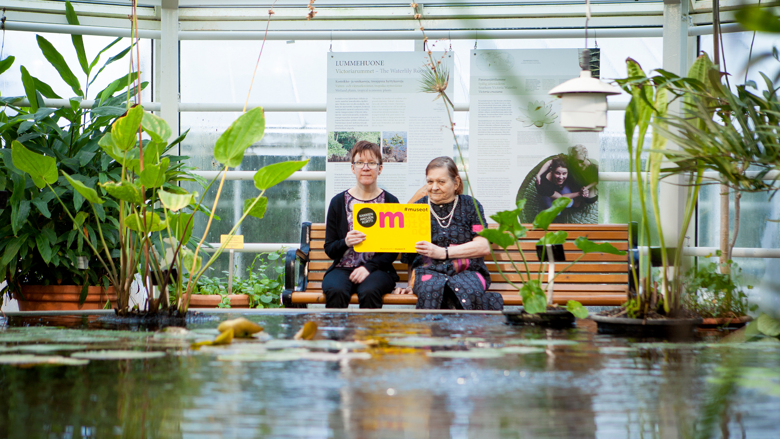 Kuvassa on kaksi naista istumassa iso museokortti kädessään ja he istuvat vesialtaan äärellä. Altaan vieressä on viherkasveja.