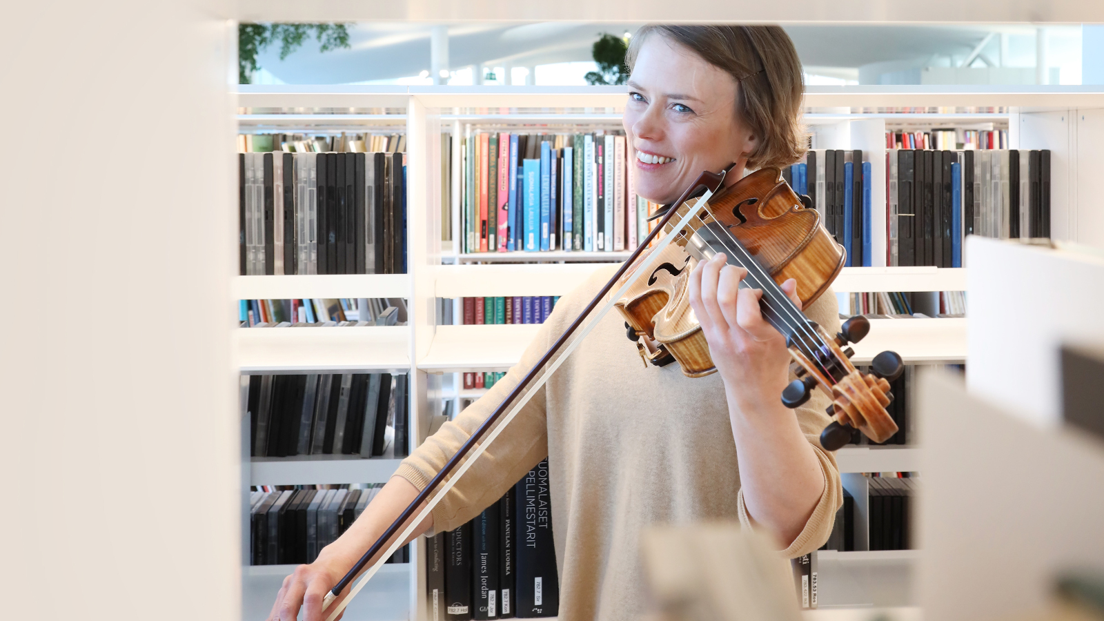 Kuvassa on nuorehko naispuolinen viulisti soittamassa kirjastossa.
