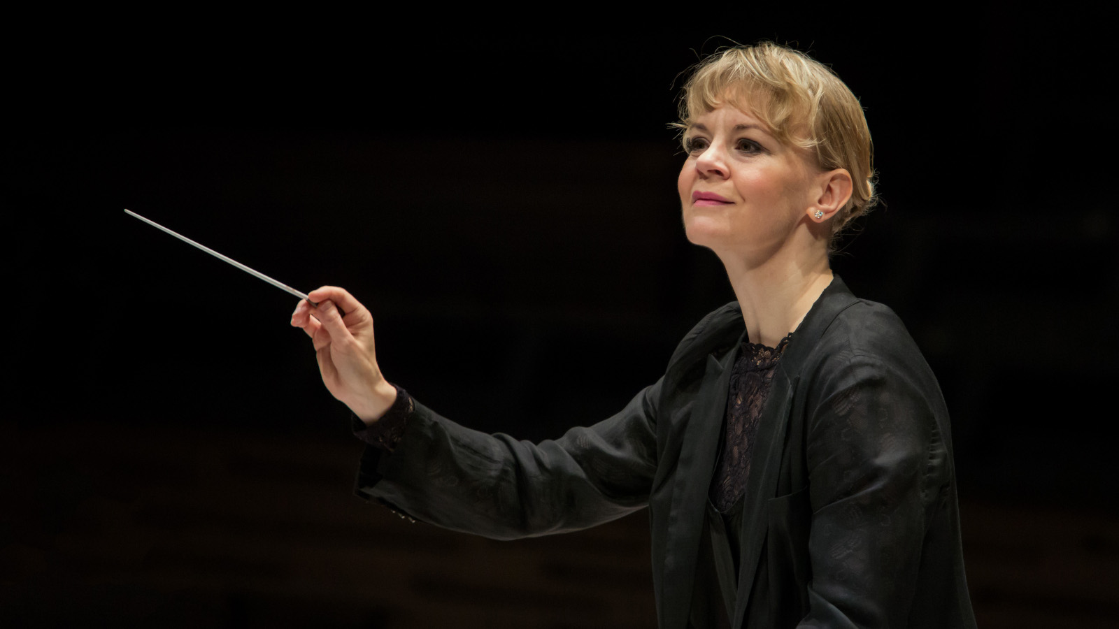 Kuvassa on Susanna Mälkki johtamassa orkesteria.  Kuva on puolivartalokuva ja Mälkillä on tahtipuikko kädessään.