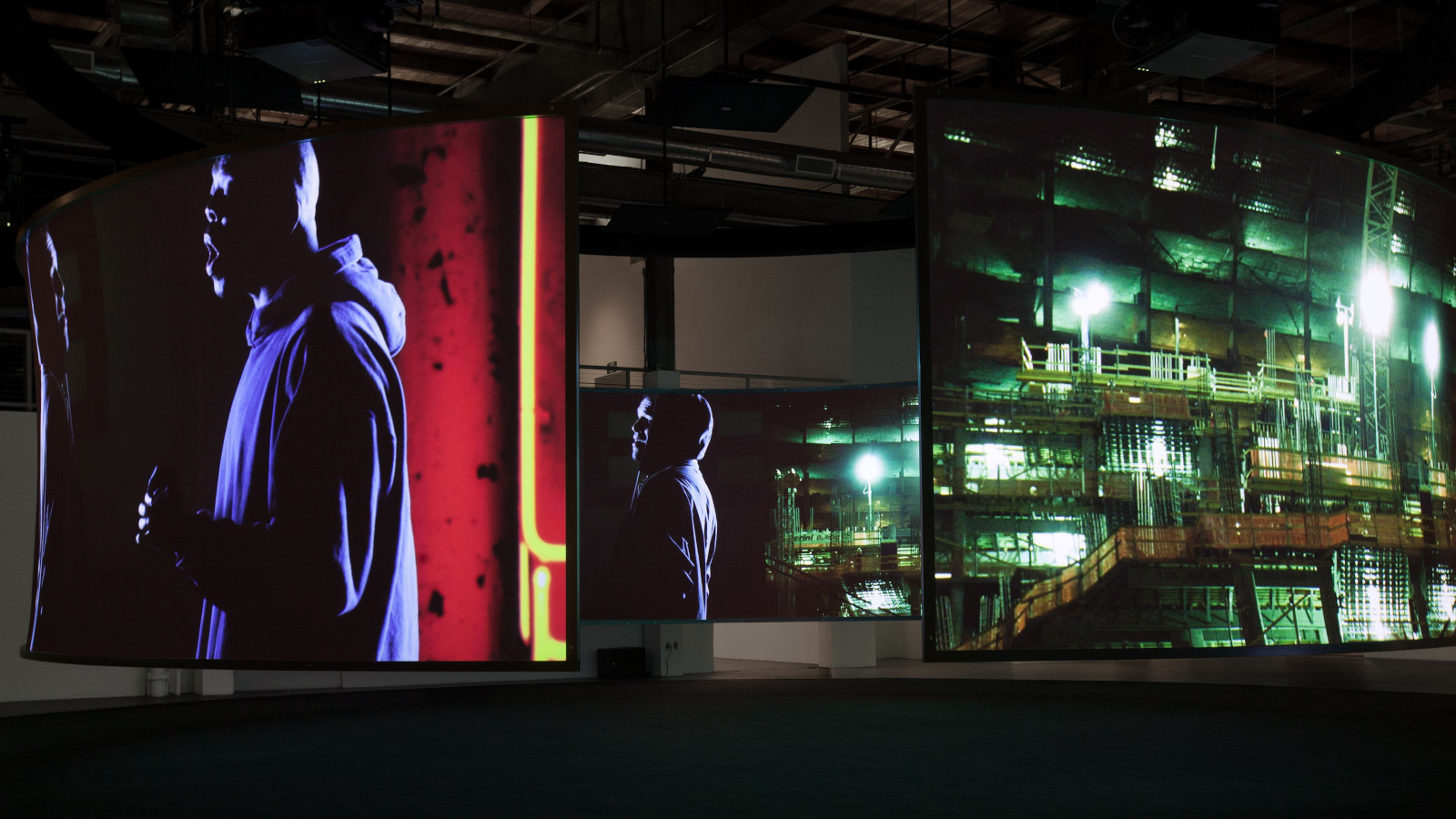 Kuvassa on Doug Aitkenin teos, jossa etualalla seisoo mies hupparissa ja vieressä on pienempi kuva miehestä. Molemmat ovat puoliksi selin. Taustalla näkyy yövalaistua modernin lasi-ikkunaisen rakennuksen seinää.