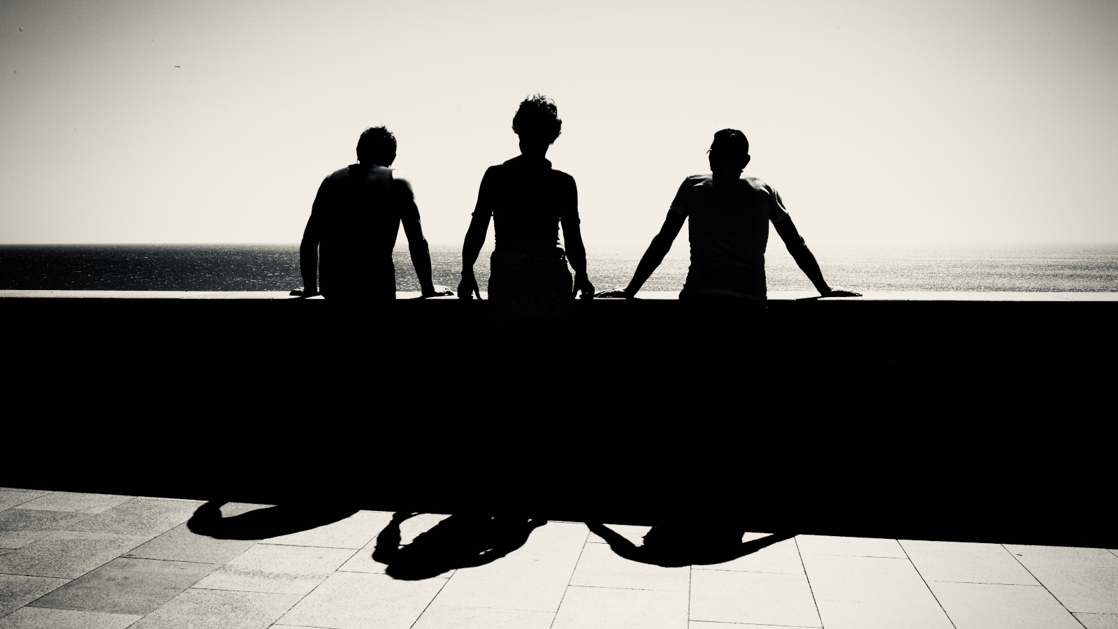 Kuvassa on kolme miestä kuvattuna takaa päin ja he katselevat merelle betonisen kaiteen äärellä. Kuva on musta-valkoinen.