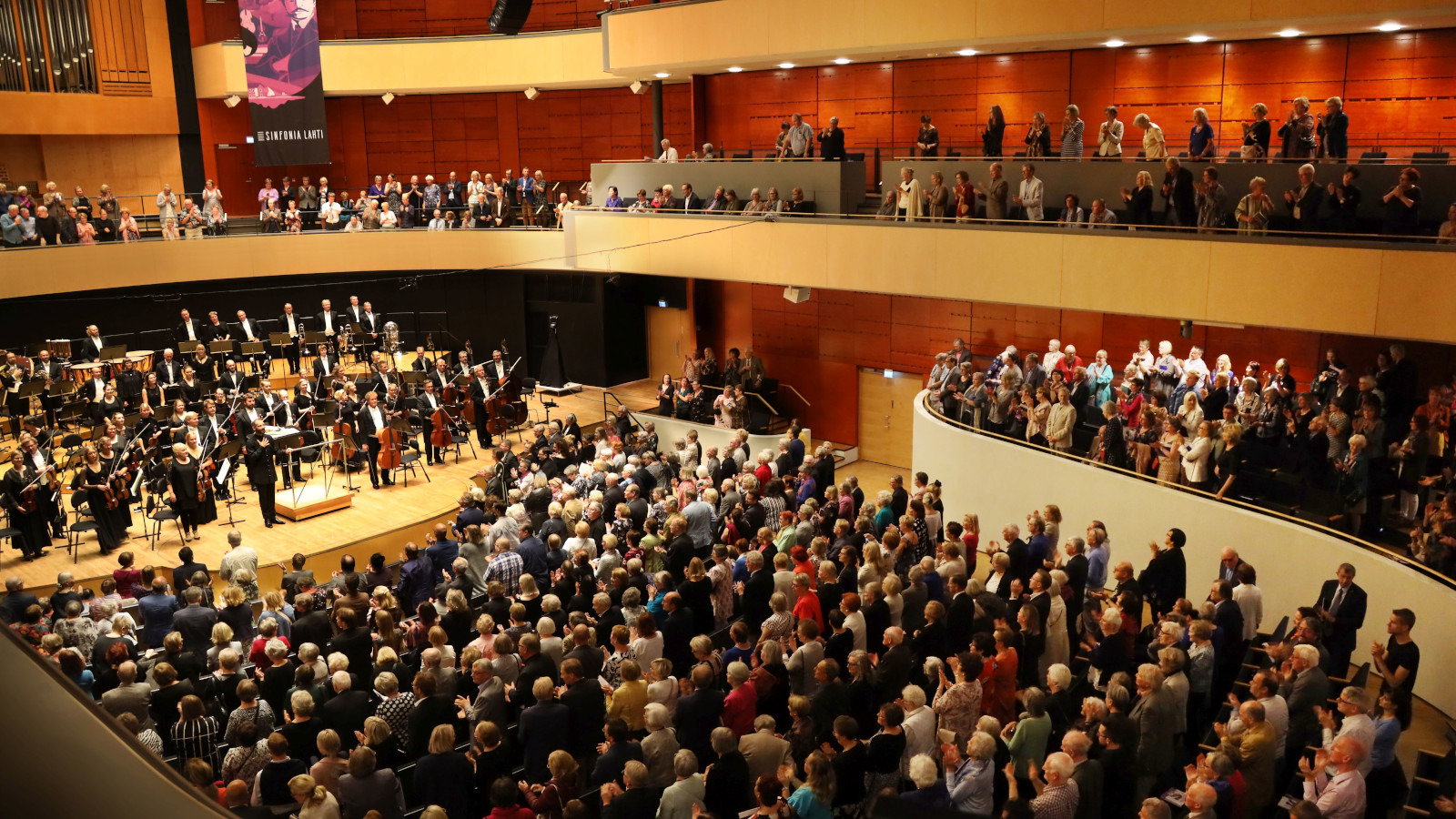 Kuva on Sibelius-talon konserttisalin konsertista, jossa on orkesteri kiittämässä ja sali täynnä yleisöä. 