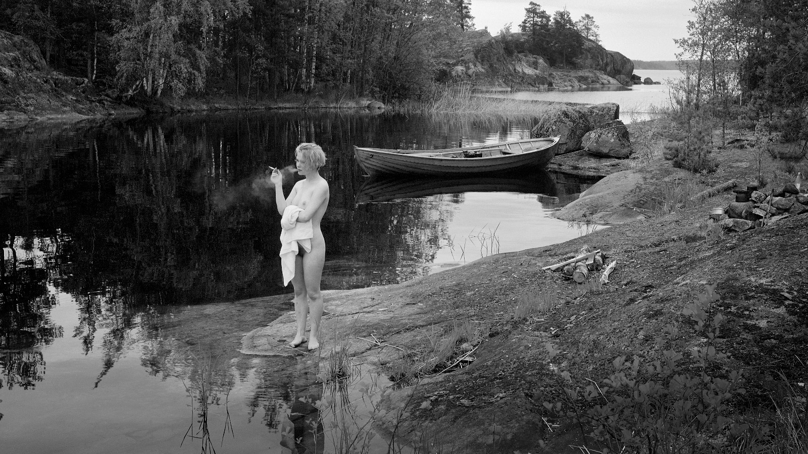 Kuvassa on nuori nainen alasti seisomassa veden äärellä matalan kallion kielekkeen päällä. 