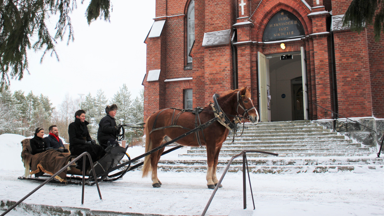 Kuvassa on hevosen vetämässä reessä Jussi Merikanto, Jussi Makkonen ja Nazig Azezian Nurmeksen punatiilisen kirkon edessä.