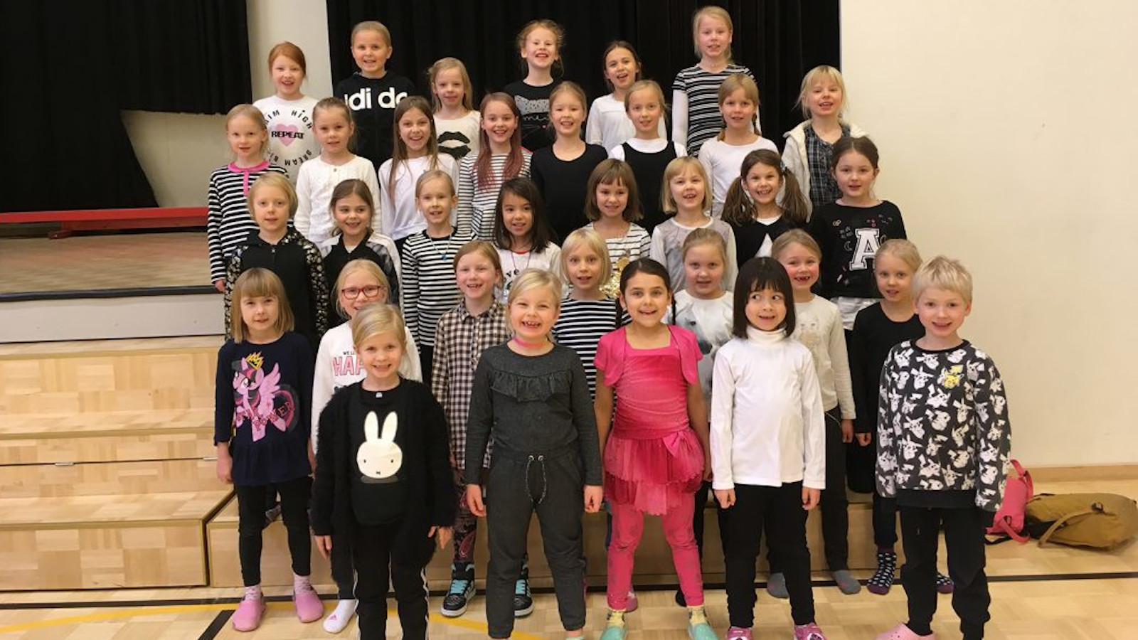 Kuvassa on Jyväskylän normaalikoulun pienten kuoro harjoituksissa ja he ovat neljässä rivissä omissa vaatteissaan.
