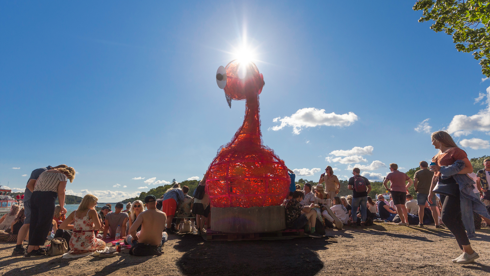 Kuvassa on Ruisrockin osallistujia istumassa hietikolla auringonpaisteessa.  Keskellä on punainen muovinen patsas, johon osuu aurinko.