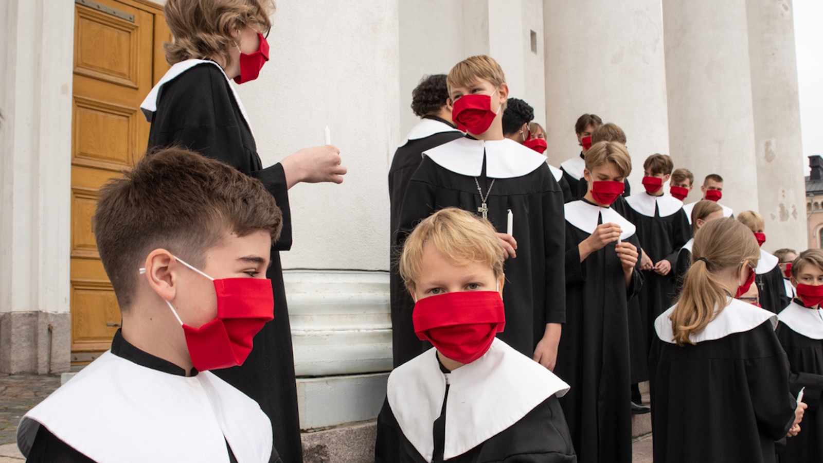Kuvassa on Cantores Minoresin nuoria laulajia mustissa kaavuissa ja punaisissa maskeissa tauolla Helsingin tuomiokirkon edessä olevassa pylväikössä.
