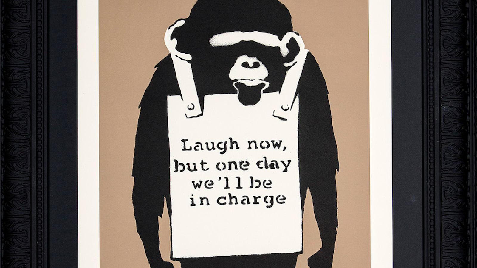 Kuvassa on osa raamitetusta taulusta, jossa on musta apina jonka rintamuksessa on taulu ja teksti 'Laugh now, but remember one day we'll be in charge'.