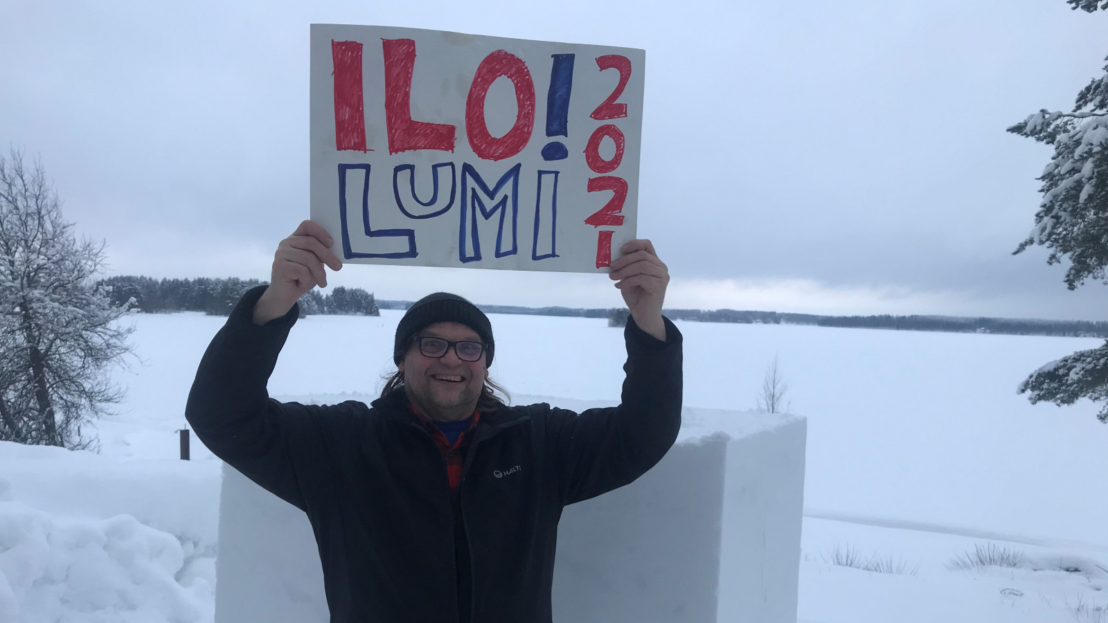 Kuvassa on Reko Mäki pitelemässä kädet ylhäällä kylttiä, jossa lukee Ilo!lumi 2021 ja taustalla on luminen järven selkä.
