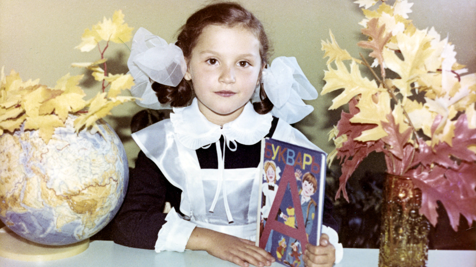 Kuvassa on Neuvostoliitossa asuva tyttö koulupuvussa isot rusetit päässa ja koulukirja kädessä.  Hän istuu valkoisen pöydän ääressä. 