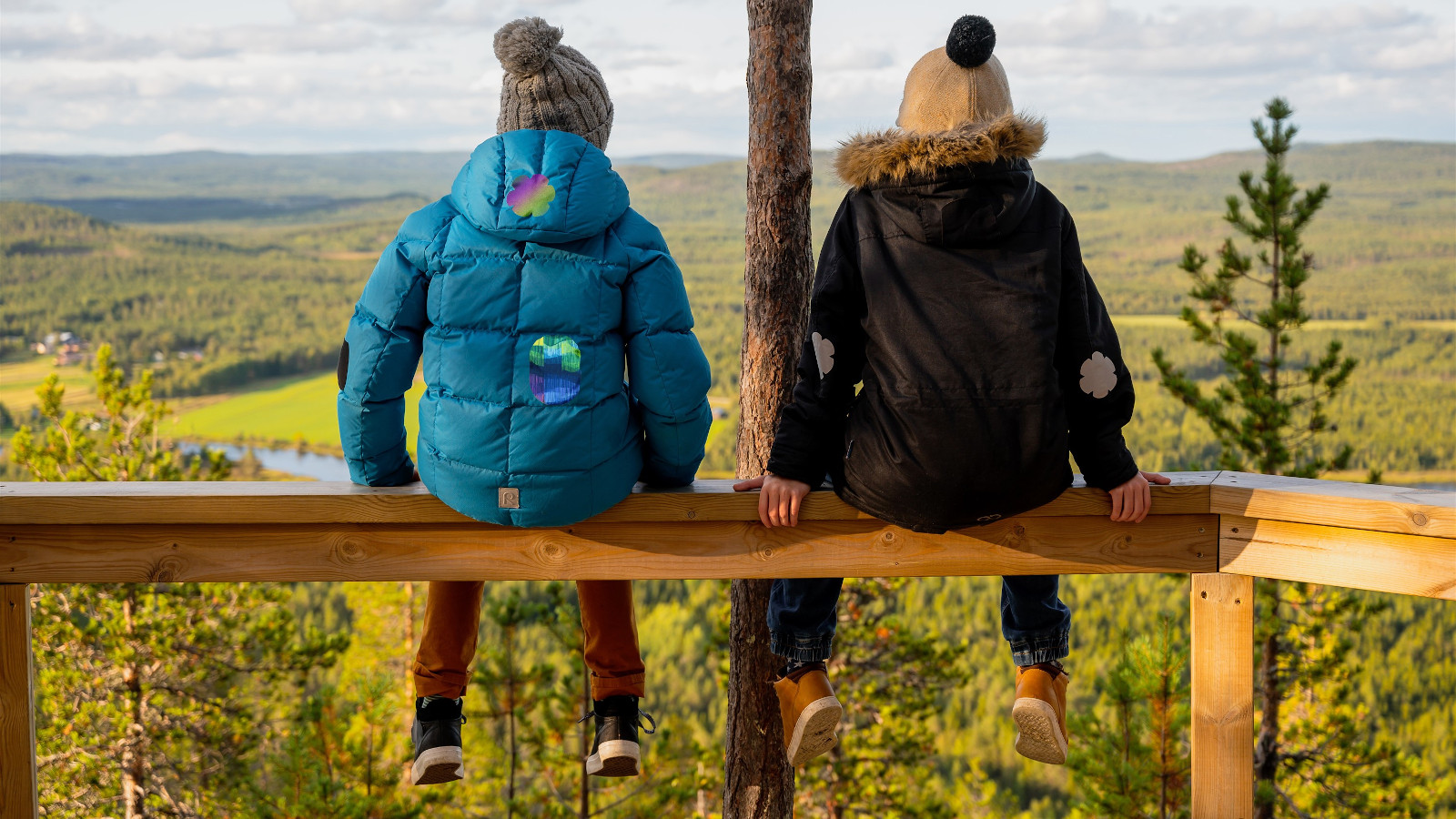 Kuvassa on kaksi tyttöä takaperin ja he istuvat aidan päällä kulmassa.  Näkymä on ylhäältä alaspäin.  Maisema on syksyisen värikäs laaja metsämaisema.